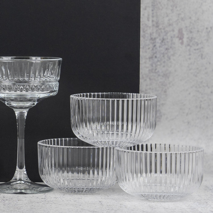 The Hamper Boutique Co’s Gin and Homeware Essentials Hamper features ribbed glass bowls and a matching stemmed glass with vertical ridges, elegantly arranged on a light surface against a black and gray background.