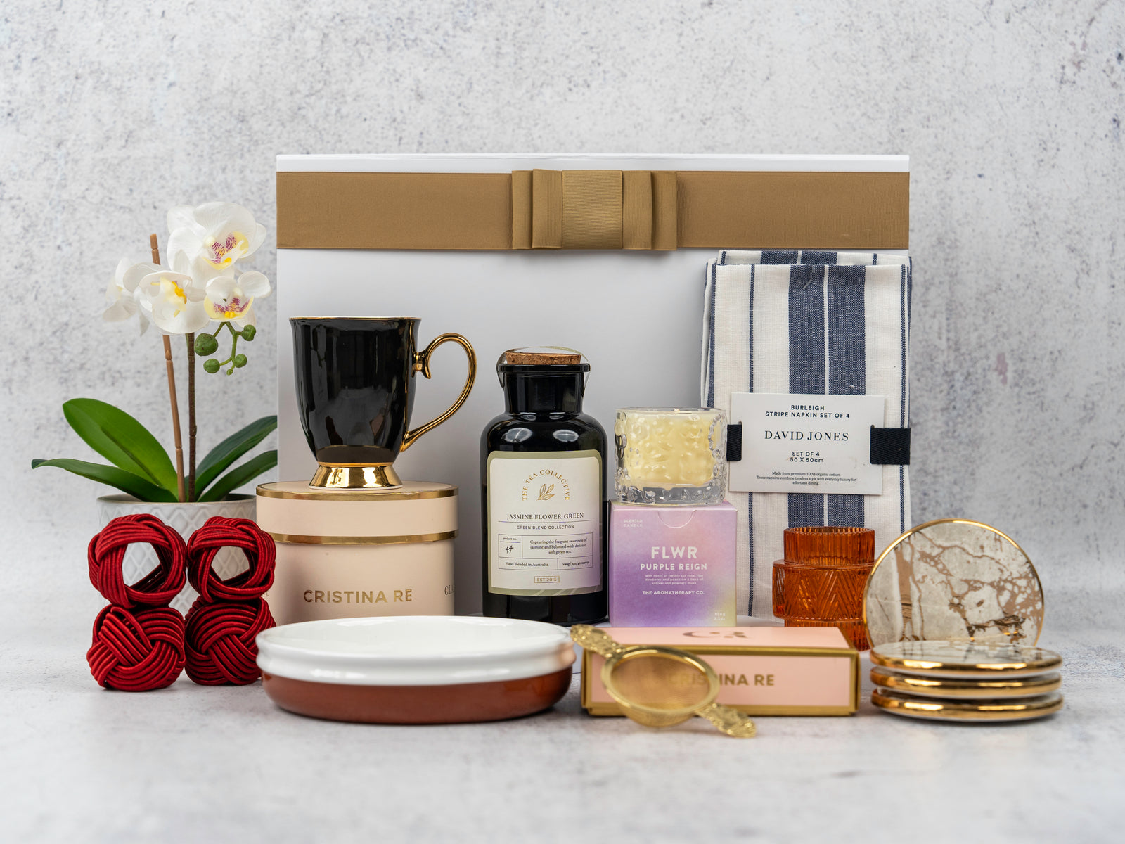 The Hamper Boutique Co's Stylish Home Entertaining Hamper is a luxurious gift set with a potted orchid, black and gold mug, syrup bottle, candle, jam, towel, napkin rings, coasters, gold tea strainer, and more—all beautifully arranged.