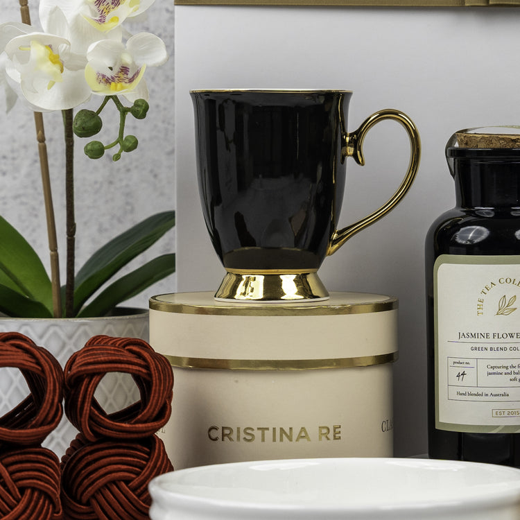 The Stylish Home Entertaining Hamper by The Hamper Boutique Co features a black and gold teacup on a round cream and gold box, paired with jasmine flower tea, a white orchid, and red woven coasters.