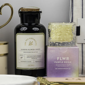 A Stylish Home Entertaining Hamper by The Hamper Boutique Co is on display, featuring Jasmine Flower Green tea beside a FLWR Purple Reign box topped with a patterned candle, arranged with other decorative accents.