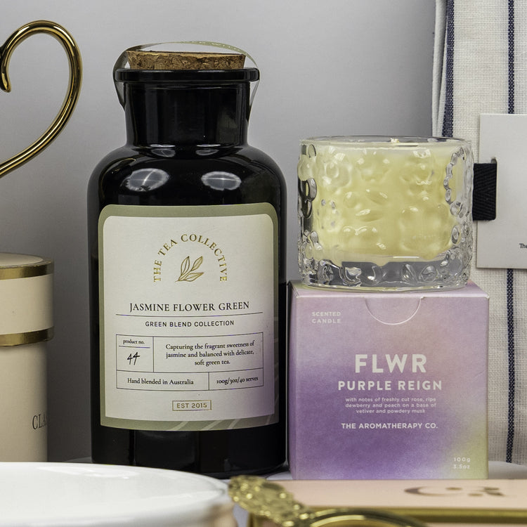 A Stylish Home Entertaining Hamper by The Hamper Boutique Co is on display, featuring Jasmine Flower Green tea beside a FLWR Purple Reign box topped with a patterned candle, arranged with other decorative accents.
