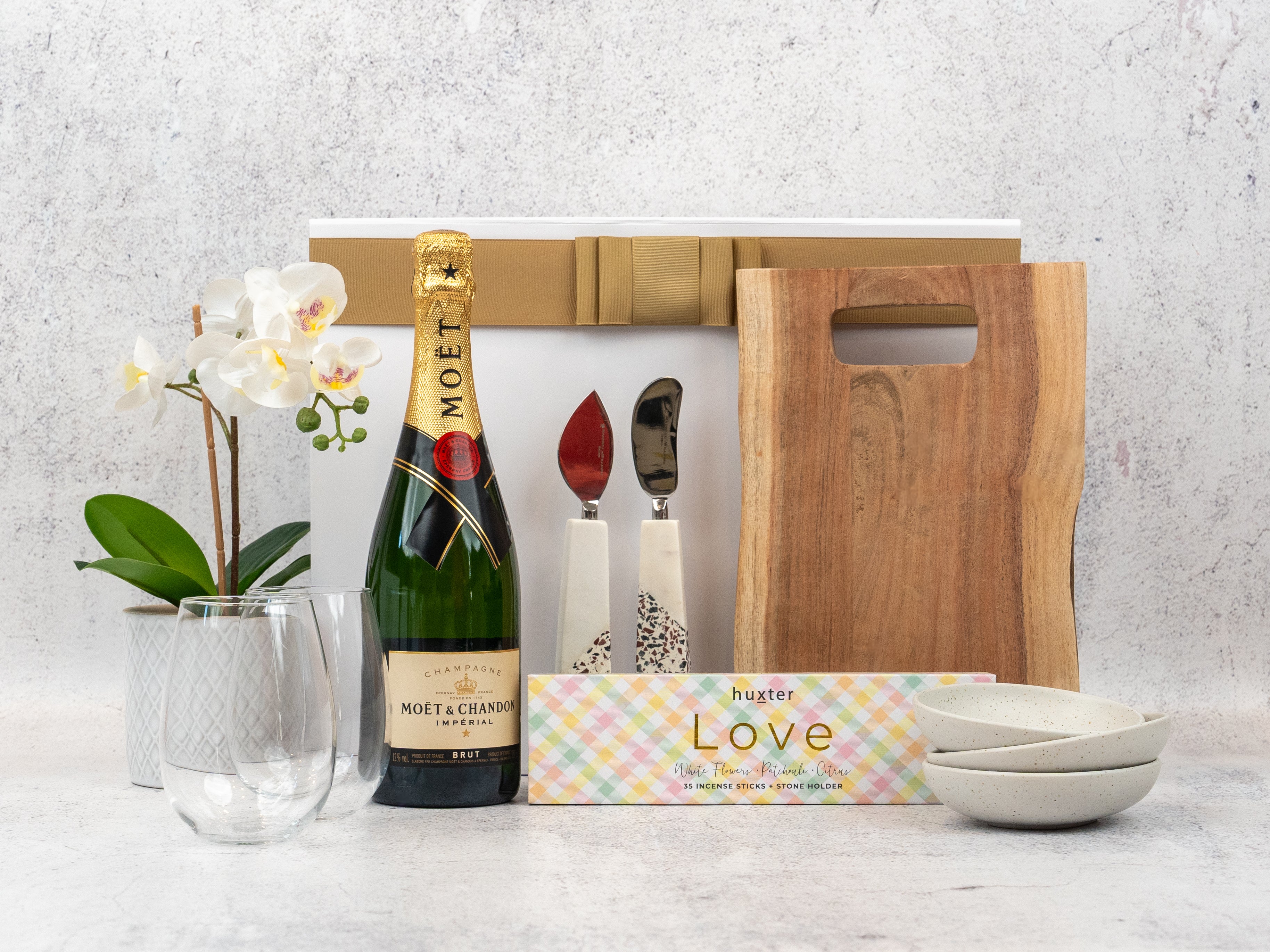 The Hamper Boutique Co’s Champagne and Homeware Collection includes Moët & Chandon, a wooden board, two ceramic bowls, marble-handled cutlery, two glasses, a white potted orchid, and a Love box—perfect for gifting.