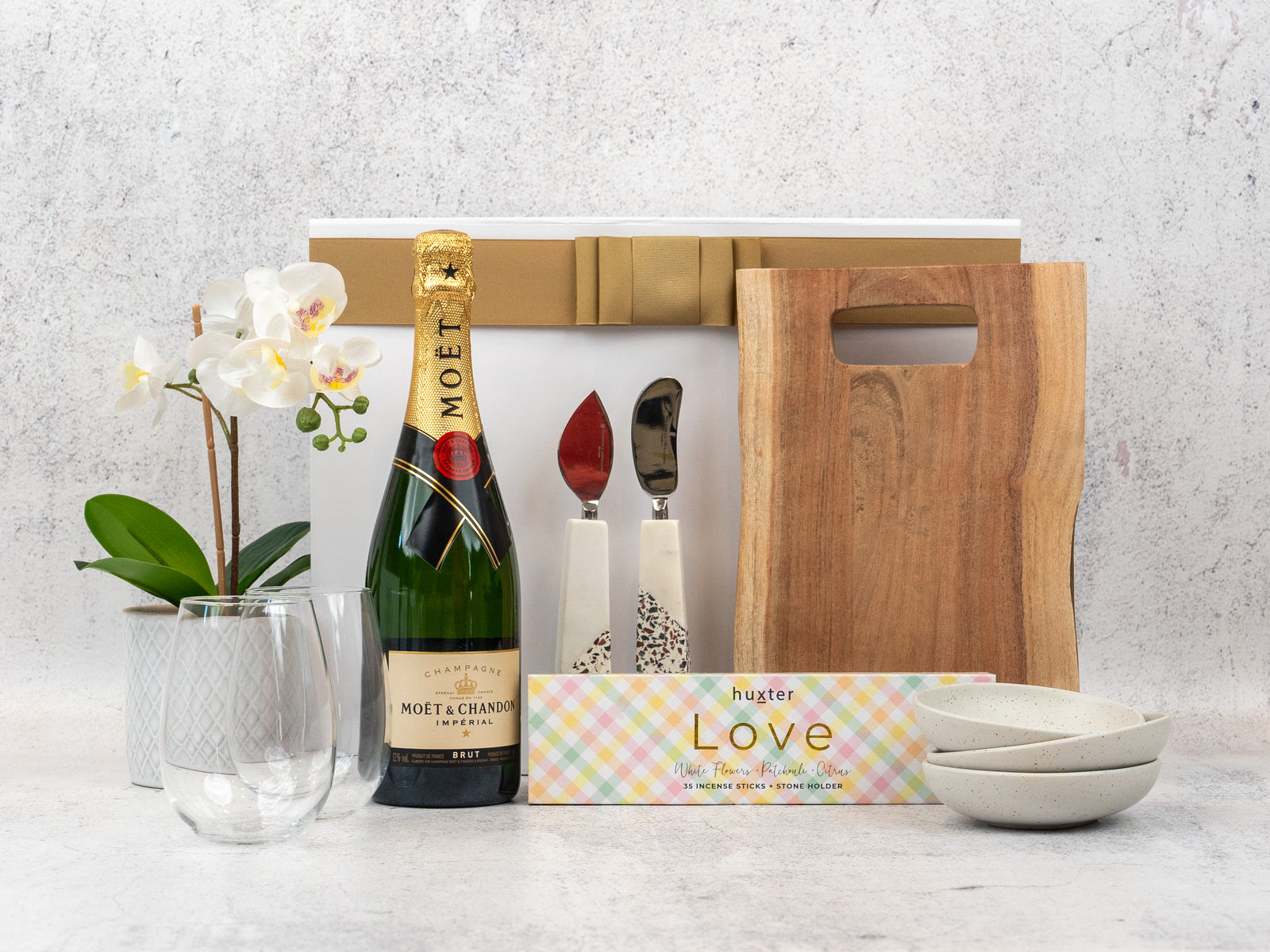 The Hamper Boutique Co’s Champagne and Homeware Collection includes Moët & Chandon, a wooden board, two ceramic bowls, marble-handled cutlery, two glasses, a white potted orchid, and a Love box—perfect for gifting.