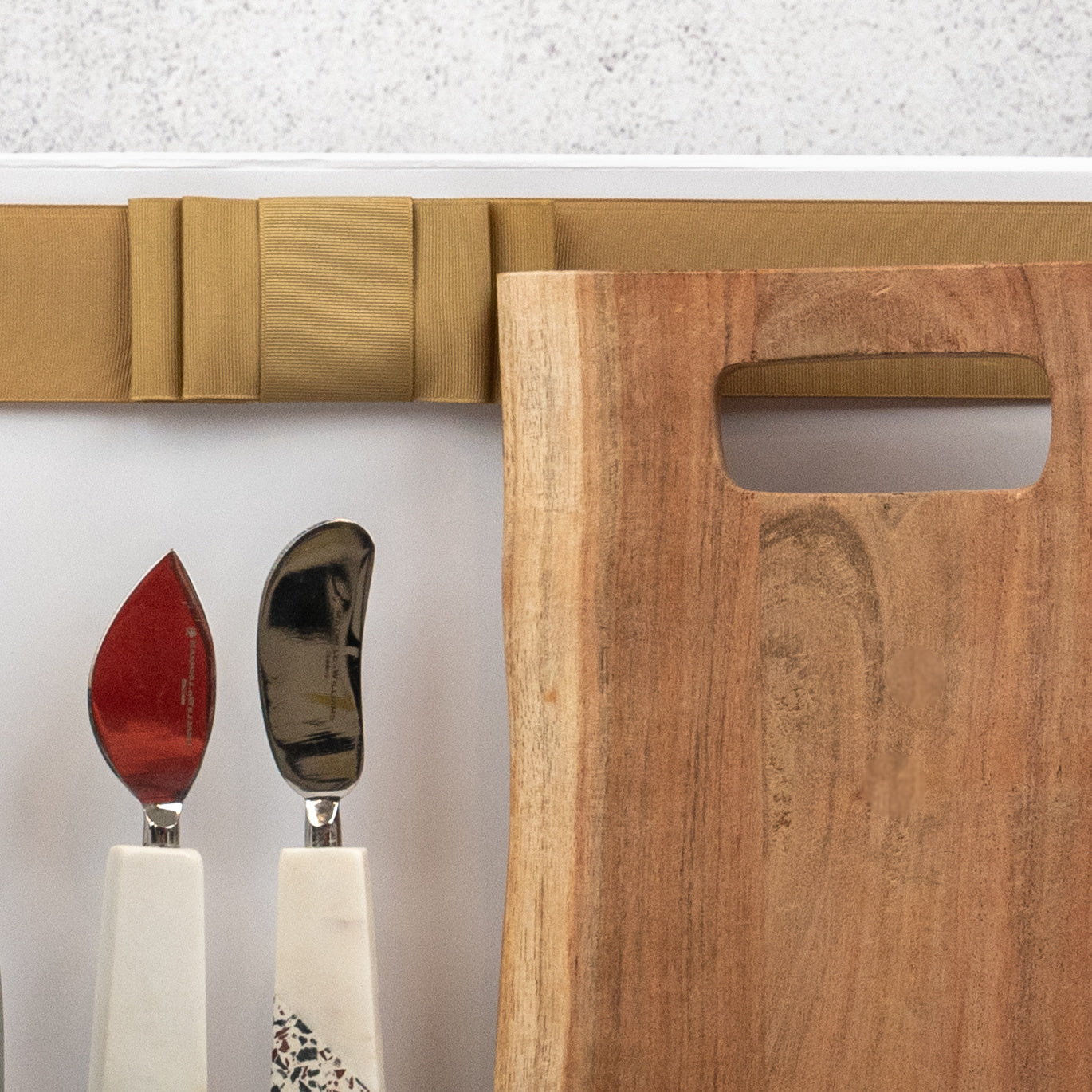 The Hamper Boutique Co's Champagne and Homeware Collection features three cheese knives with white handles and a wooden cutting board, elegantly arranged on a white surface with a folded beige napkin above.