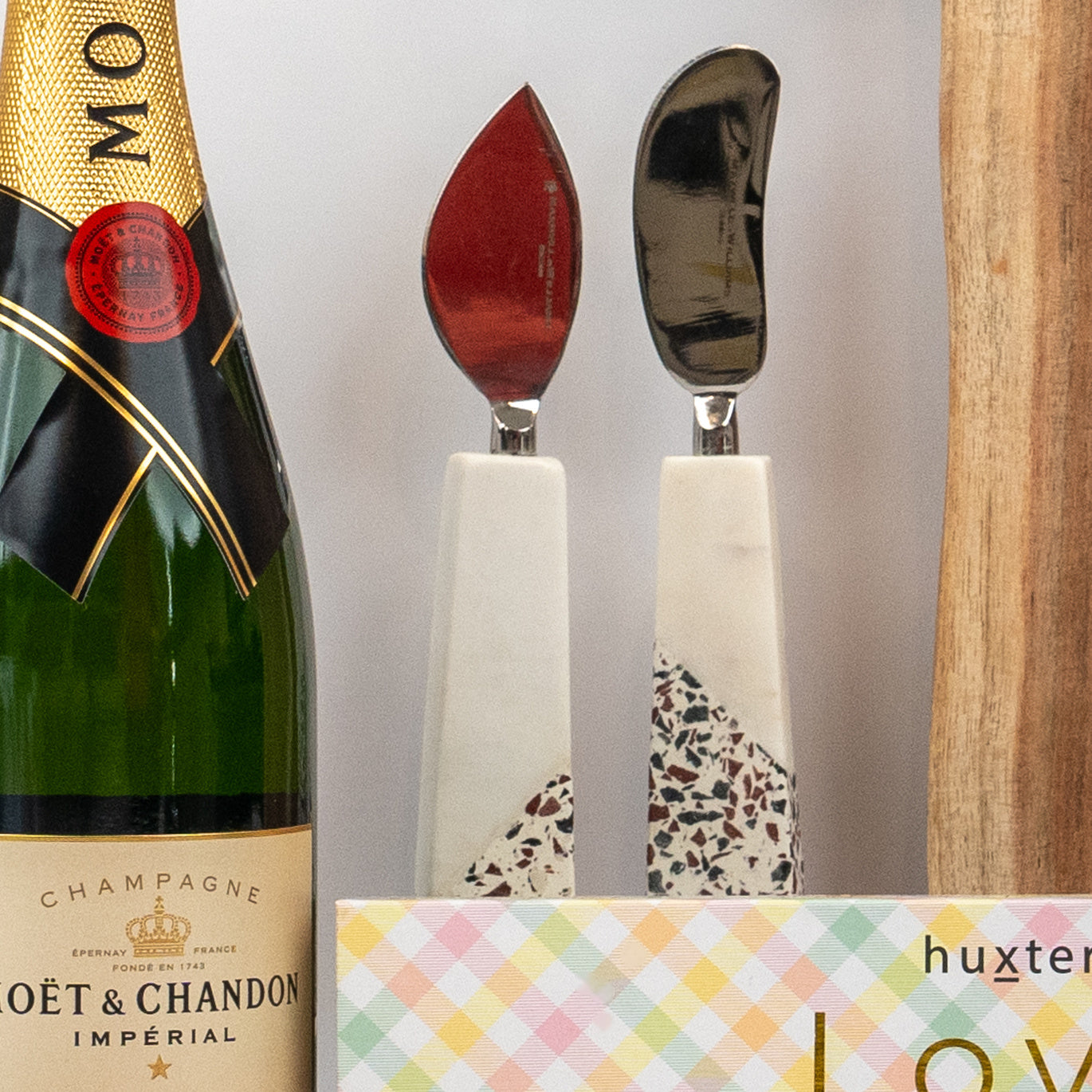 A bottle from The Hamper Boutique Co’s Champagne and Homeware Collection sits beside terrazzo cheese knives, a pastel box, and a wooden board.