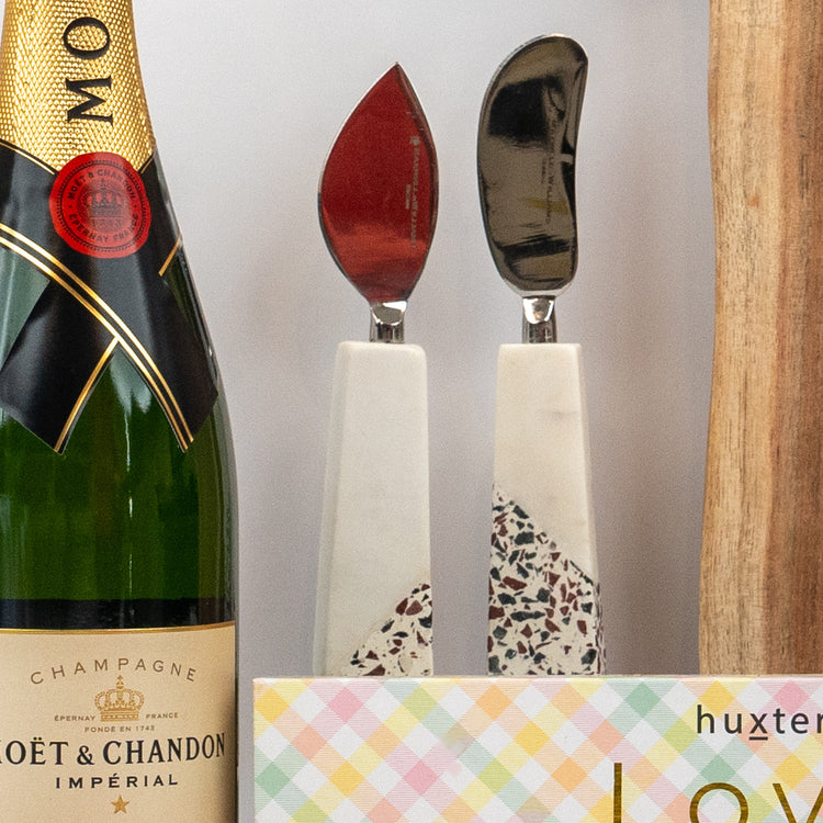 A bottle from The Hamper Boutique Co’s Champagne and Homeware Collection sits beside terrazzo cheese knives, a pastel box, and a wooden board.