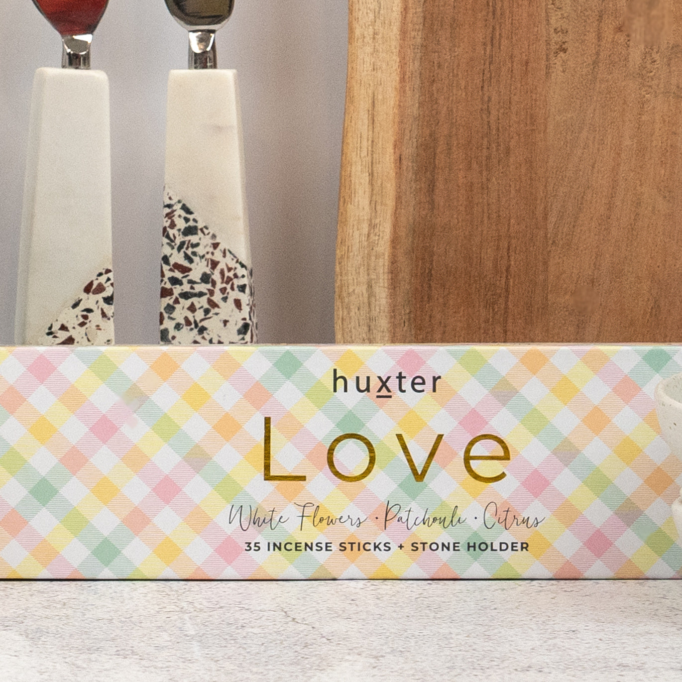 A pastel plaid box from The Hamper Boutique Co’s Champagne and Homeware Collection holds 35 incense sticks with a stone holder on a light surface, with decorative utensils and a wooden cutting board in the background.