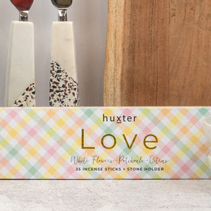 A pastel plaid box from The Hamper Boutique Co’s Champagne and Homeware Collection holds 35 incense sticks with a stone holder on a light surface, with decorative utensils and a wooden cutting board in the background.