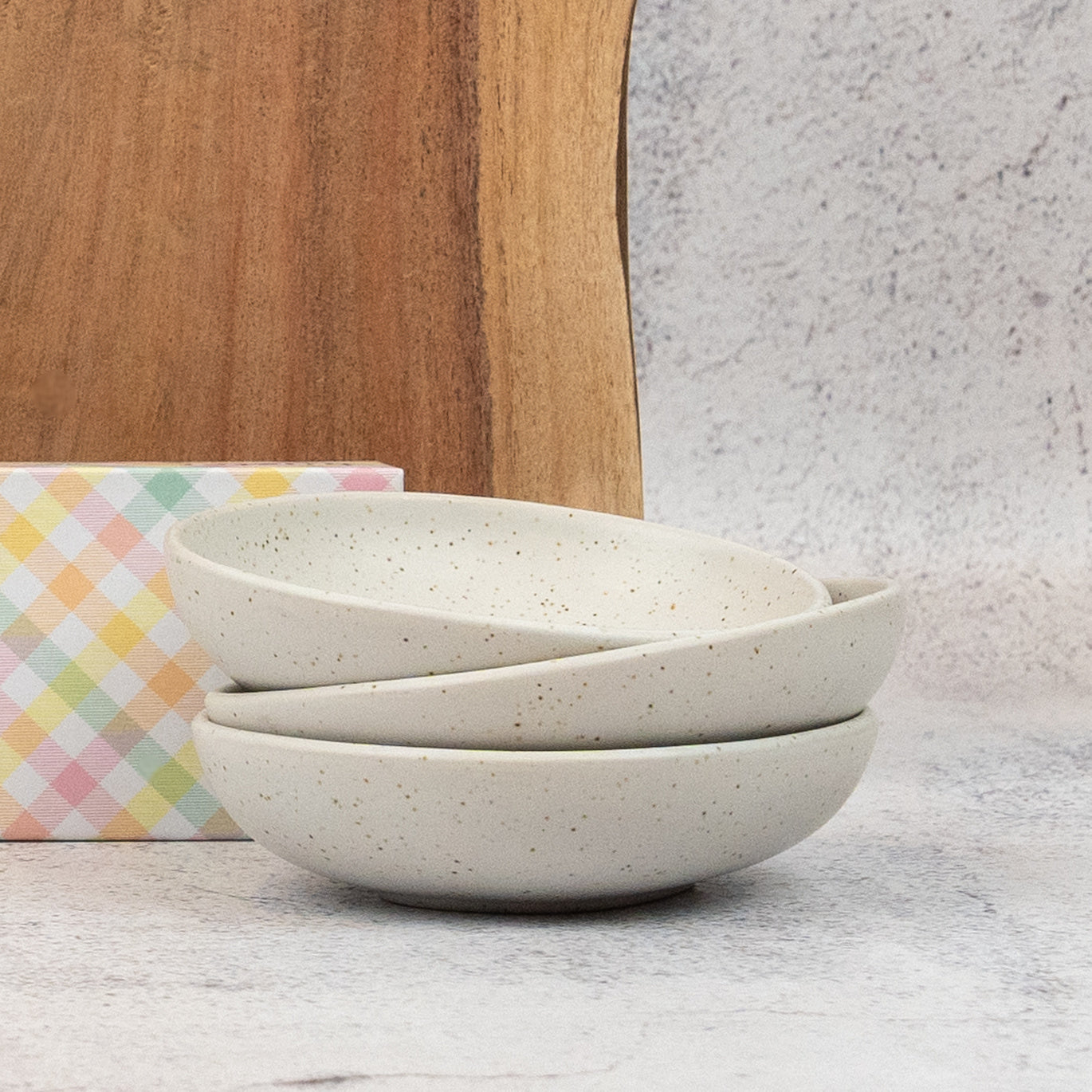 Three stacked speckled ceramic bowls from The Hamper Boutique Co’s Champagne and Homeware Collection are displayed in front of a pastel checkered box and wooden cutting board against a light, textured background.