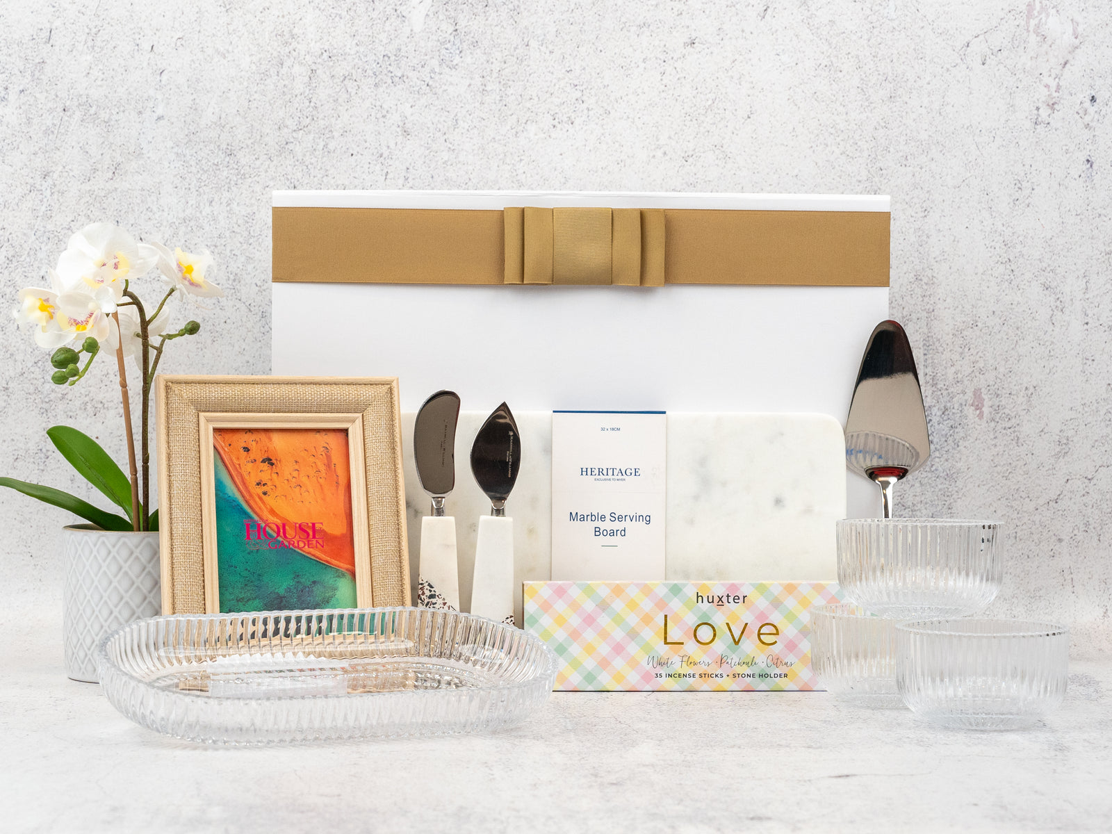 The Hamper Boutique Co’s Homeware Bliss Gift Hamper features a gold-ribboned box, marble board, gold utensils, framed “HOUSE” print, two glass bowls, glass tray, potted white orchid, and “Love” soap bar—all beautifully arranged.