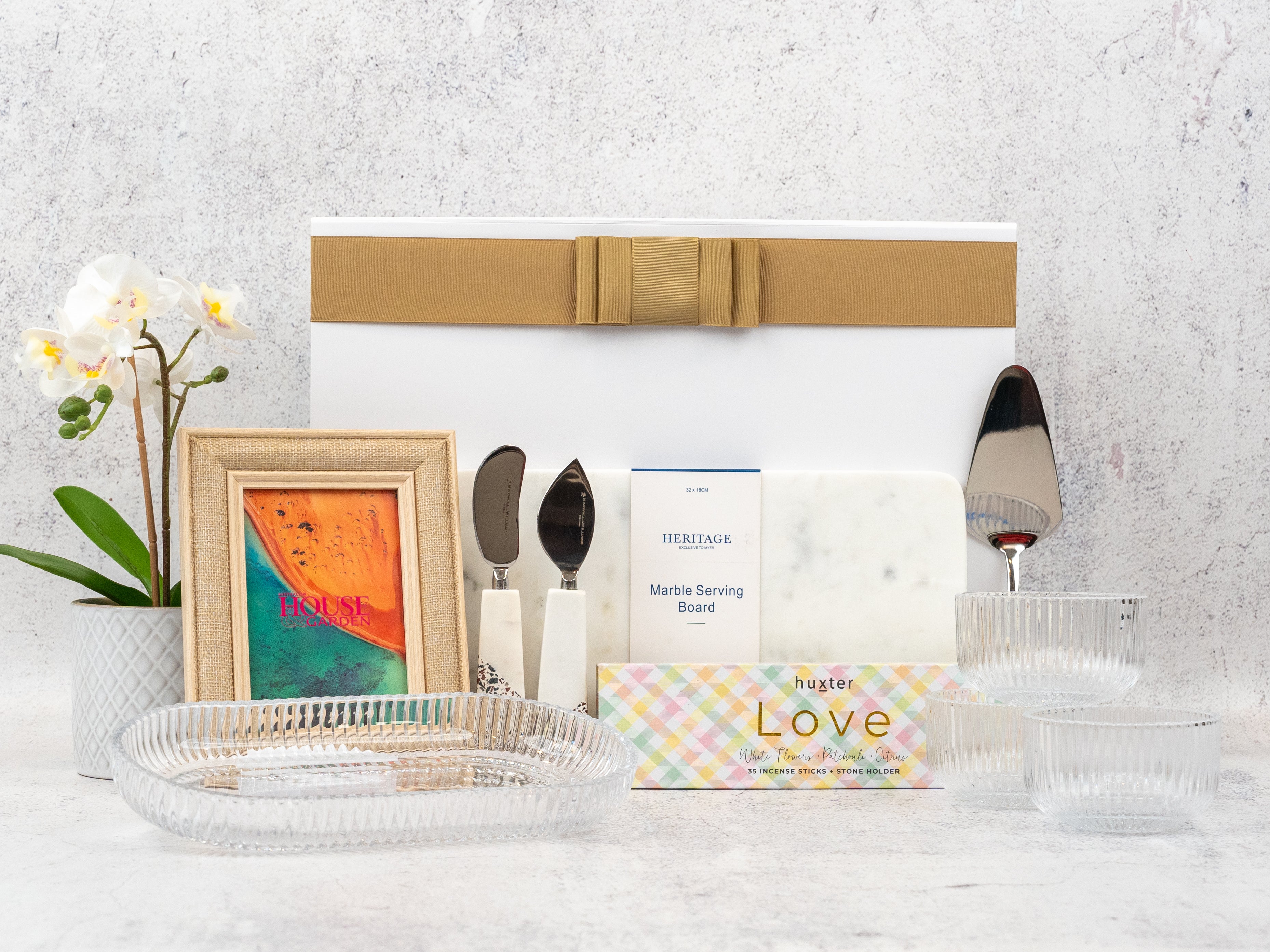 The Hamper Boutique Co’s Homeware Bliss Gift Hamper features a gold-ribboned box, marble board, gold utensils, framed “HOUSE” print, two glass bowls, glass tray, potted white orchid, and “Love” soap bar—all beautifully arranged.
