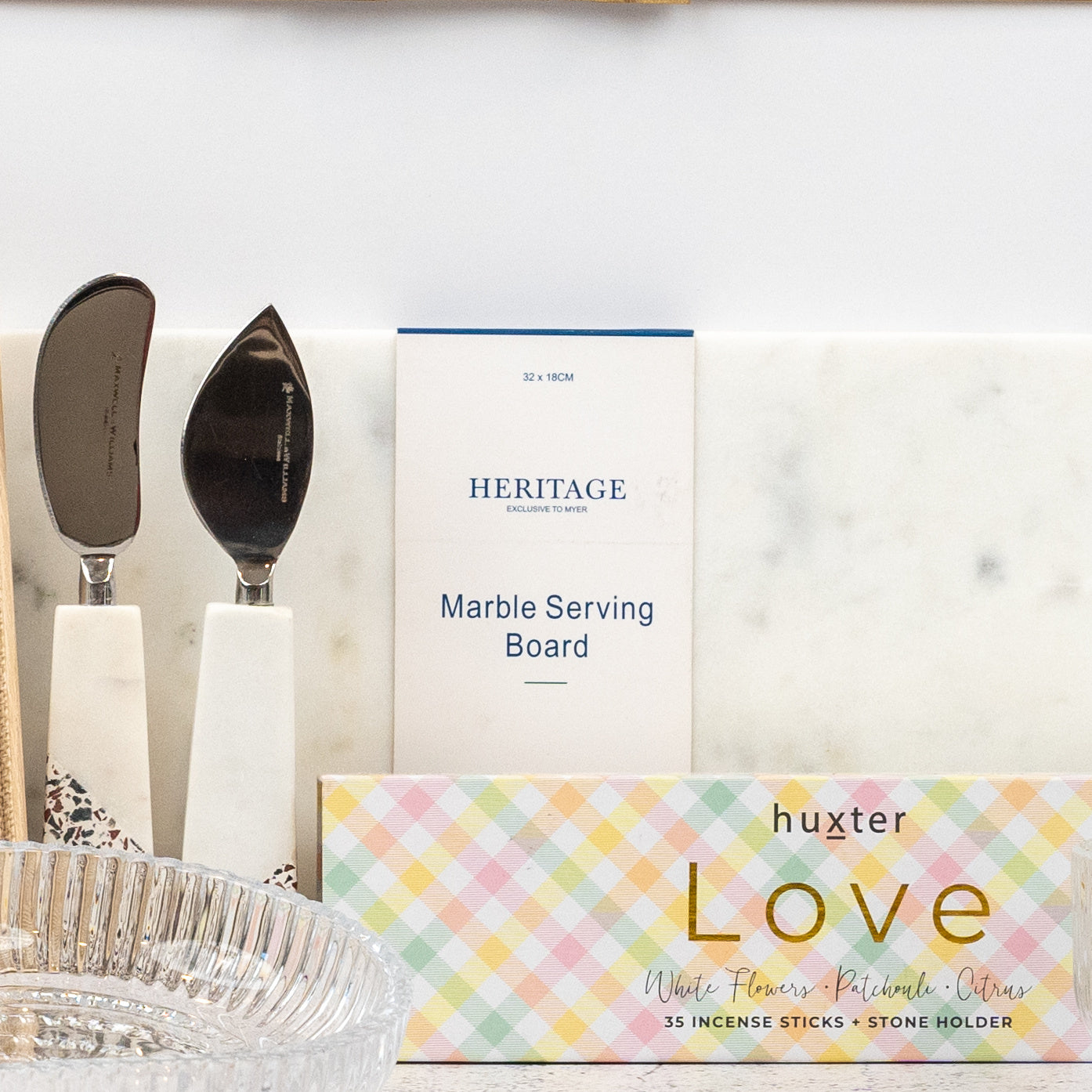 A close-up of the Homeware Bliss Gift Hamper by The Hamper Boutique Co on a counter, featuring cheese knives, a marble serving board, a glass dish, and a pastel box of huxter Love incense sticks.