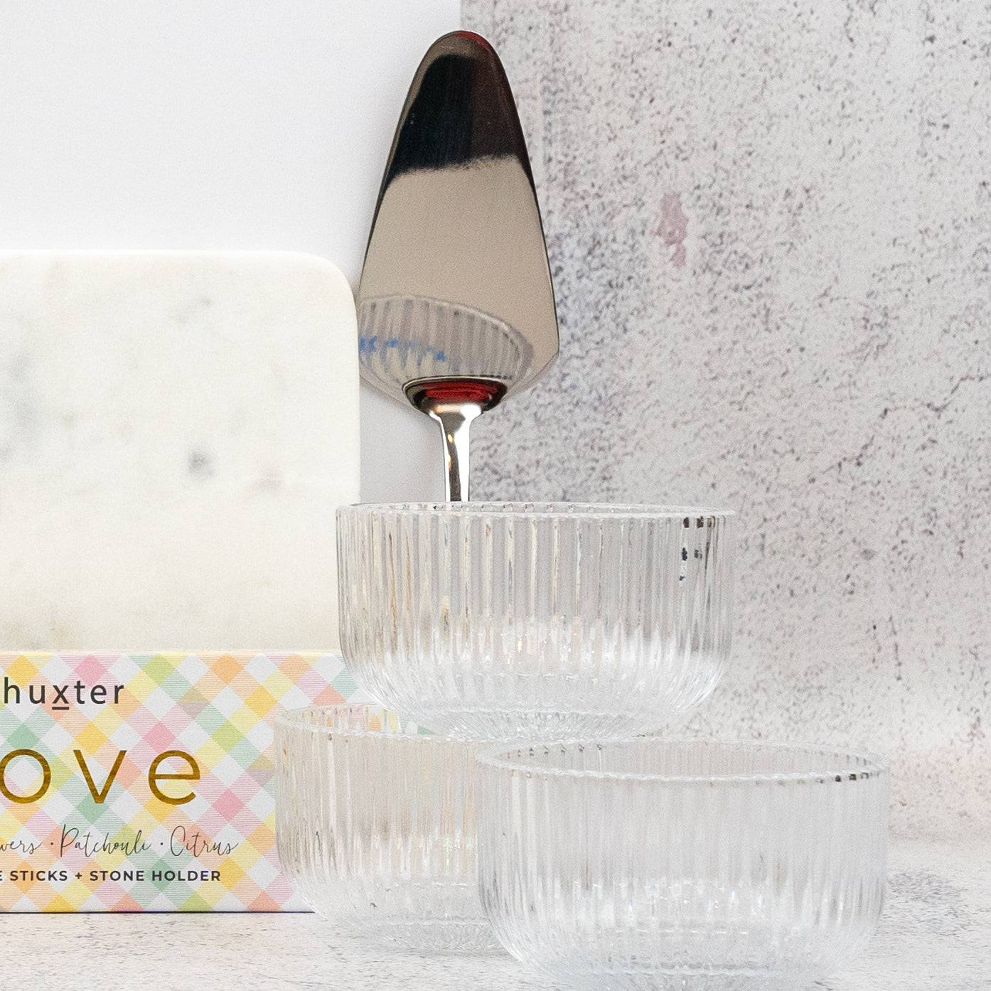 Three clear, ribbed glass bowls from The Hamper Boutique Co's Homeware Bliss Gift Hamper sit in front of a marble cheese board, cake server, and pastel plaid "Love" box on a light textured surface.
