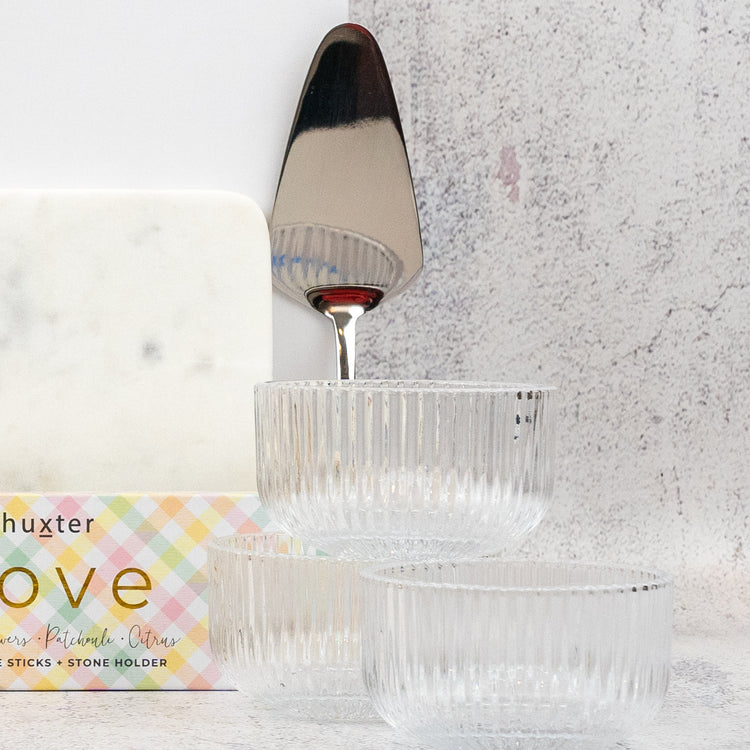 Three clear, ribbed glass bowls from The Hamper Boutique Co's Homeware Bliss Gift Hamper sit in front of a marble cheese board, cake server, and pastel plaid "Love" box on a light textured surface.