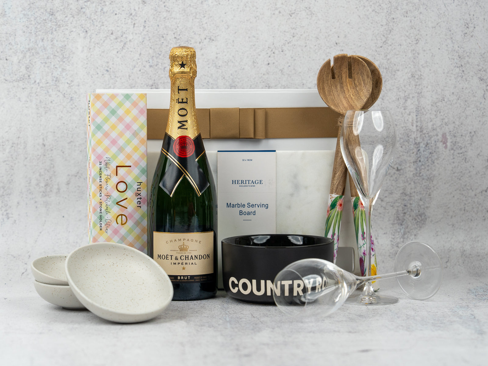 The Hamper Boutique Co Ultimate Celebration Hamper includes Moët & Chandon champagne, two champagne flutes, nougat, a marble serving board, two white bowls, a black COUNTRY bowl, and wooden salad servers.