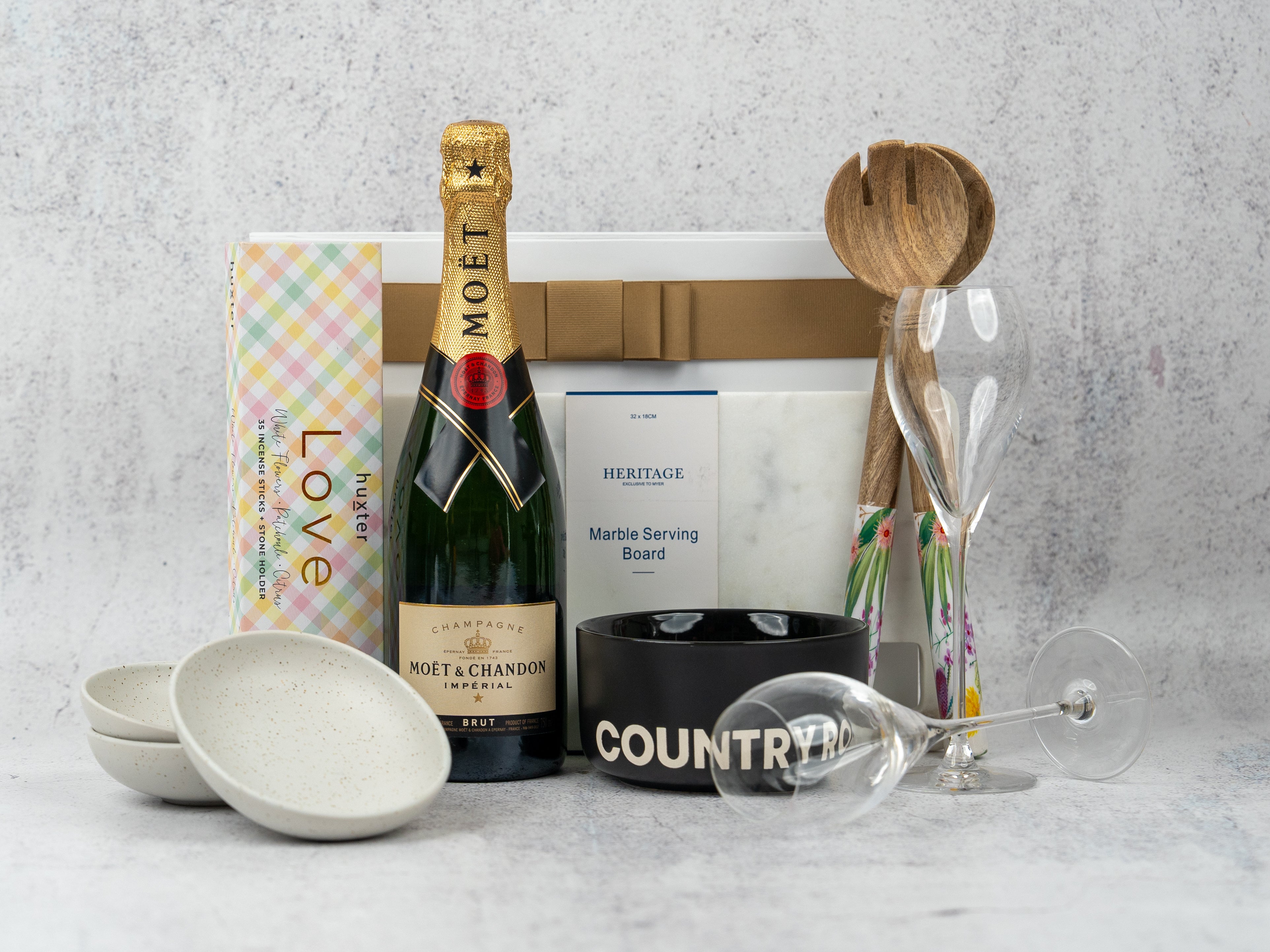 The Hamper Boutique Co Ultimate Celebration Hamper includes Moët & Chandon champagne, two champagne flutes, nougat, a marble serving board, two white bowls, a black COUNTRY bowl, and wooden salad servers.
