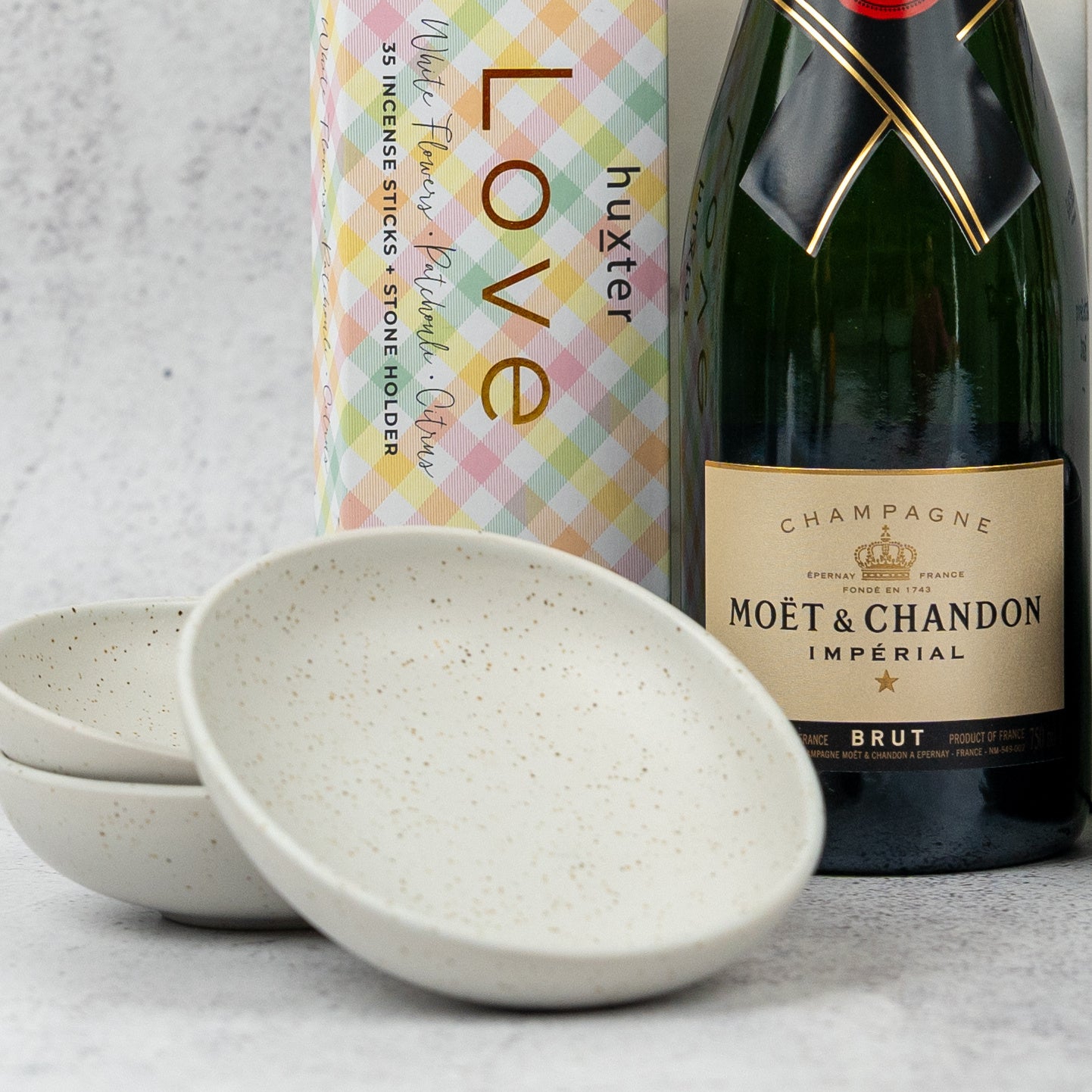 An Ultimate Celebration Hamper from The Hamper Boutique Co sits beside a colorful boxed incense set labeled Love, with three stacked speckled ceramic bowls arranged in front on a light gray surface.