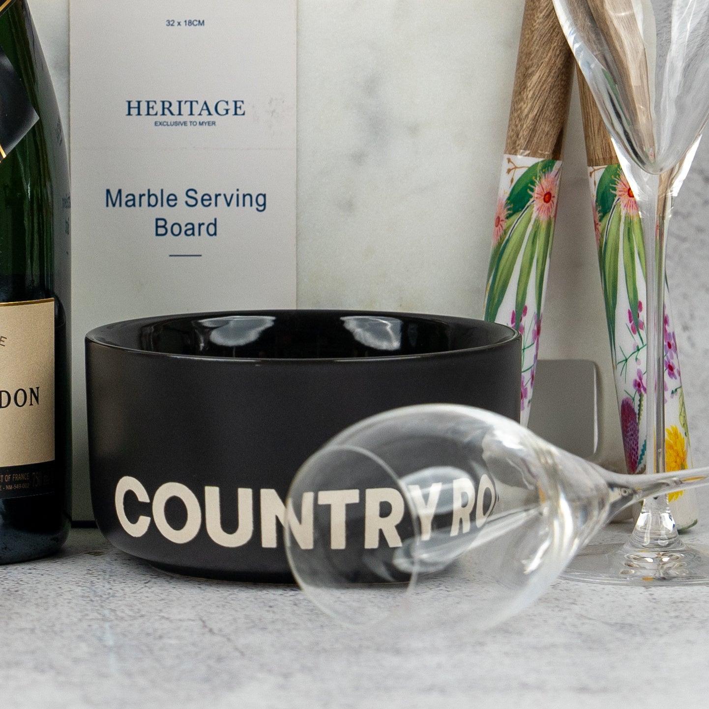 The Hamper Boutique Co's Ultimate Celebration Hamper features a black COUNTRY ROAD bowl, empty wine glass, bottle, champagne flutes, floral utensils, and a marble serving board arranged on a light surface.