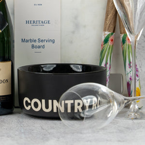 The Hamper Boutique Co's Ultimate Celebration Hamper features a black COUNTRY ROAD bowl, empty wine glass, bottle, champagne flutes, floral utensils, and a marble serving board arranged on a light surface.