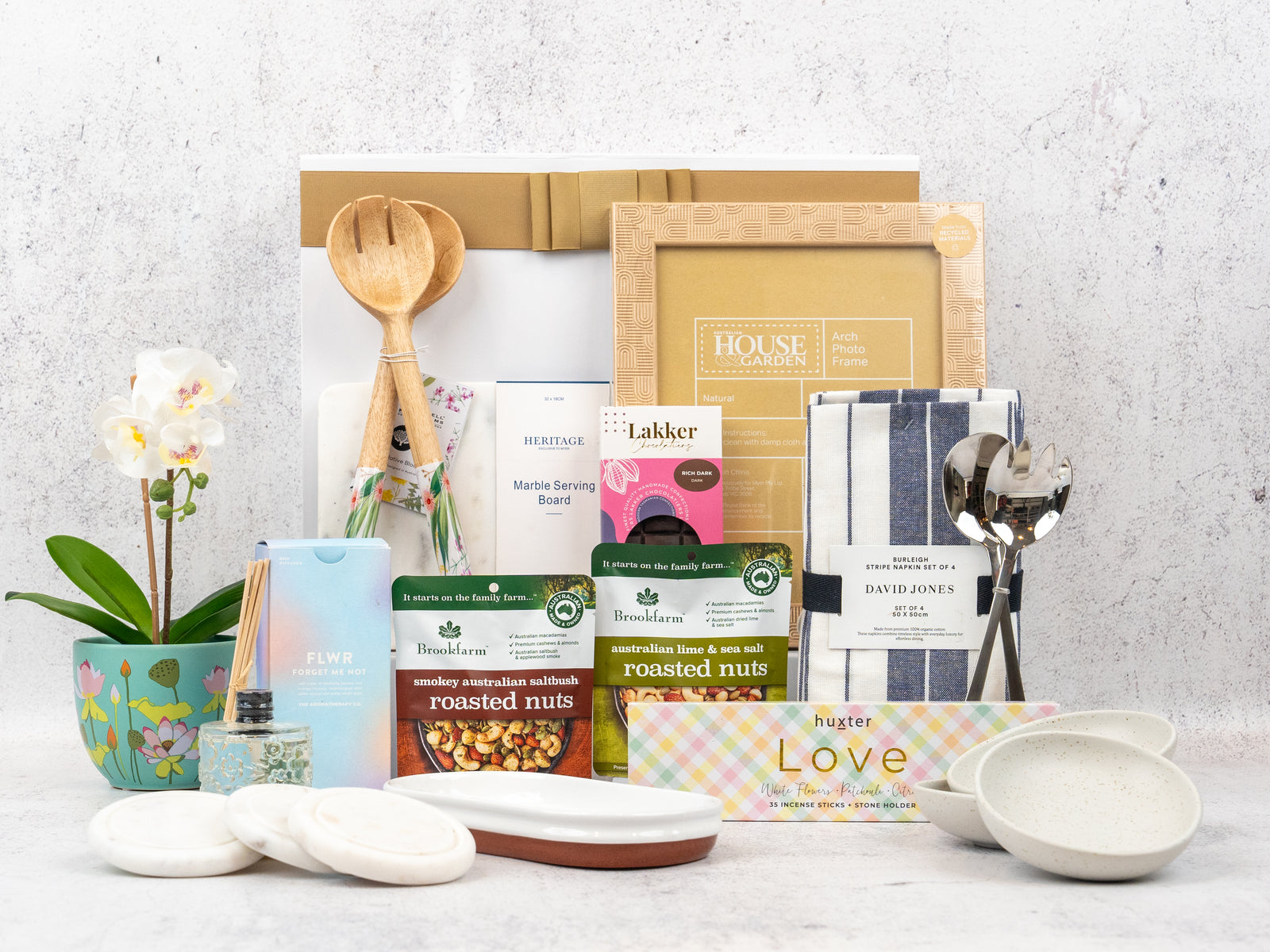 The Hamper Boutique Co's Wellness and Homeware Hamper features roasted nuts, plates, a striped tea towel, a floral mug, wooden utensils, small bowls, a serving board, a framed print, and a potted white orchid.