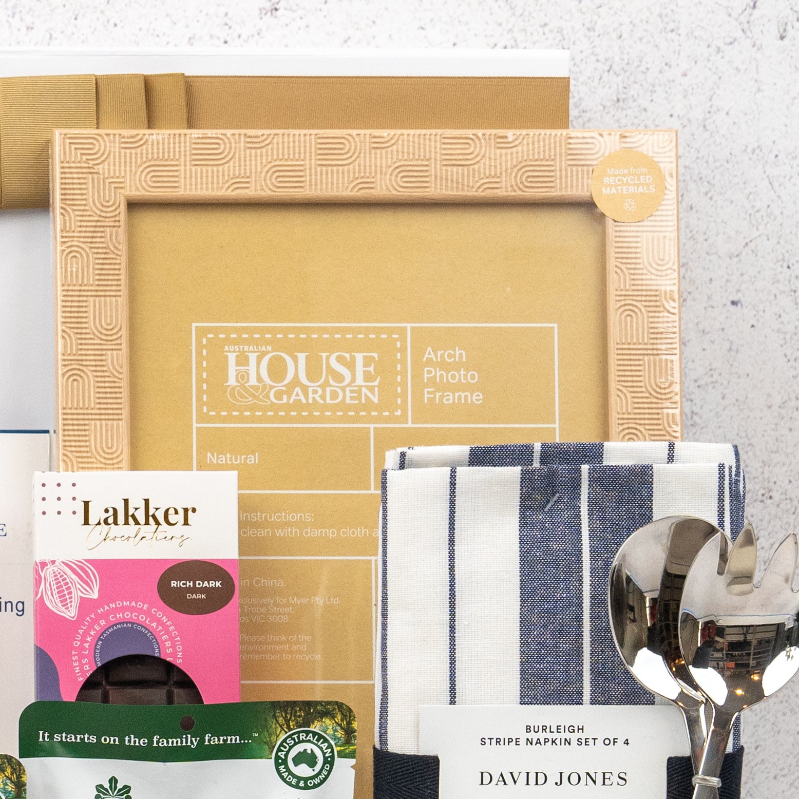 A close-up of The Hamper Boutique Co's Wellness and Homeware Hamper, featuring a picture frame, striped napkins, assorted chocolates, a green tea box, and two silver spoons, all neatly arranged against a white background.