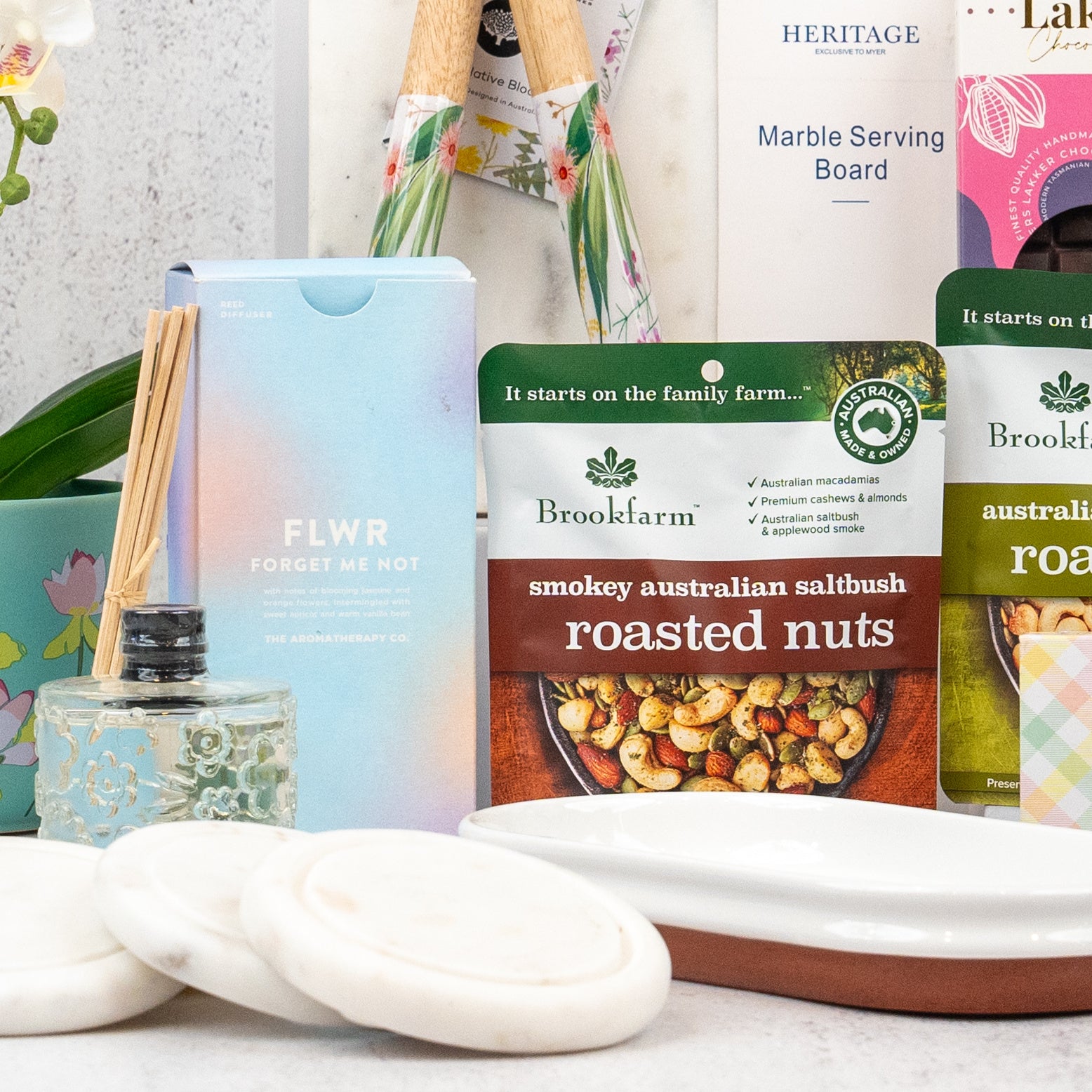 The Hamper Boutique Co’s Wellness and Homeware Hamper features roasted nuts, a FLWR forget-me-not diffuser, white round soaps, a marble serving board, and floral-themed gifts beautifully arranged on a white surface.