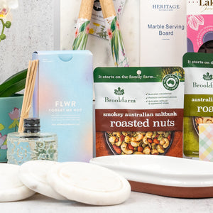 The Hamper Boutique Co’s Wellness and Homeware Hamper features roasted nuts, a FLWR forget-me-not diffuser, white round soaps, a marble serving board, and floral-themed gifts beautifully arranged on a white surface.