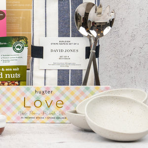 The Hamper Boutique Co’s Wellness and Homeware Hamper features a striped napkin set, serving utensils, three nested white speckled bowls, a box of nuts, and pastel-checkered incense sticks with a stone holder.