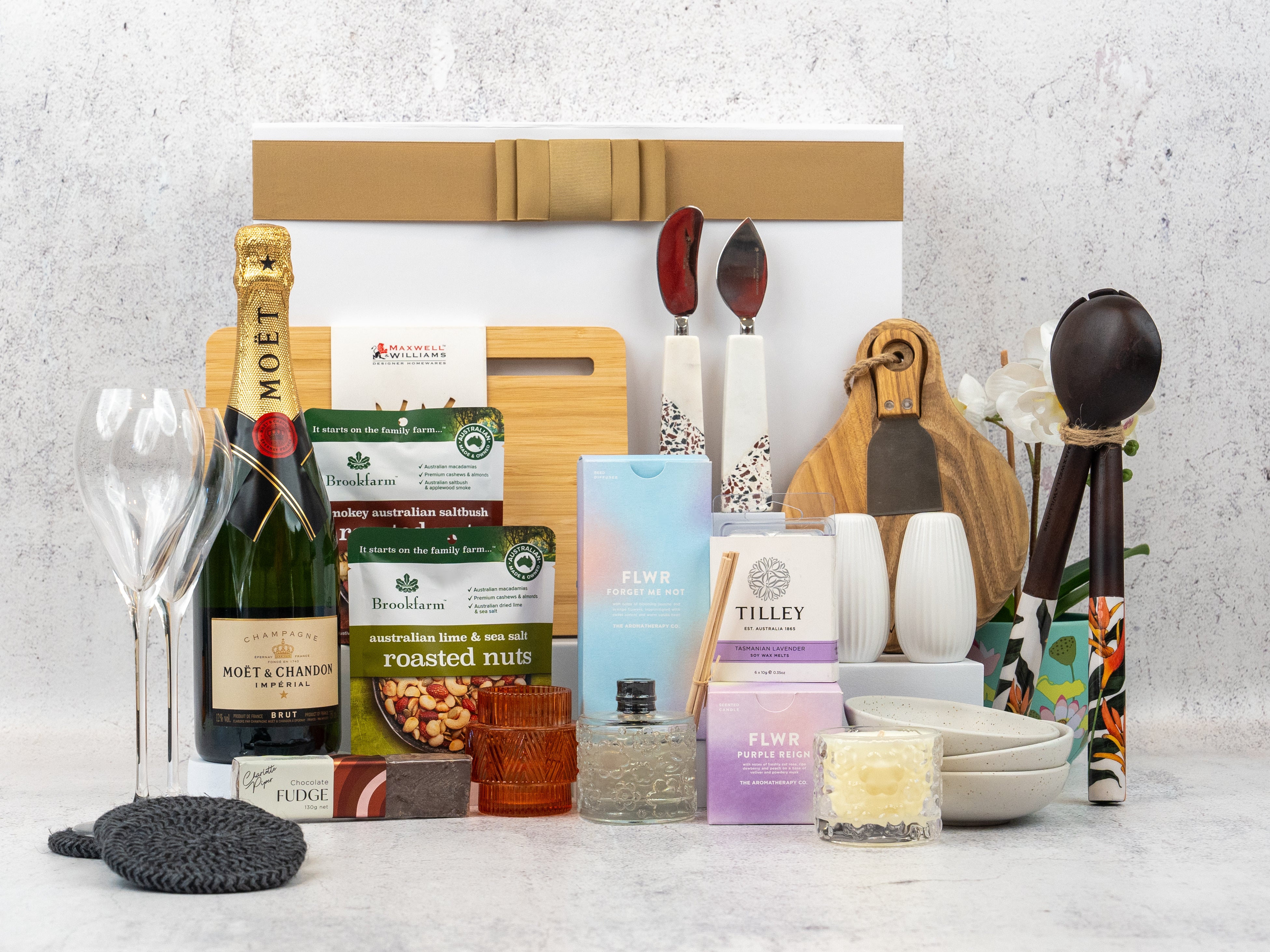 The Hamper Boutique Co’s Ultimate Homeware Gift Hamper features Moët & Chandon champagne, flutes, snacks, candles, fudge, nuts, utensils, a plant, bowls and cutting boards—beautifully arranged in front of a gift box.