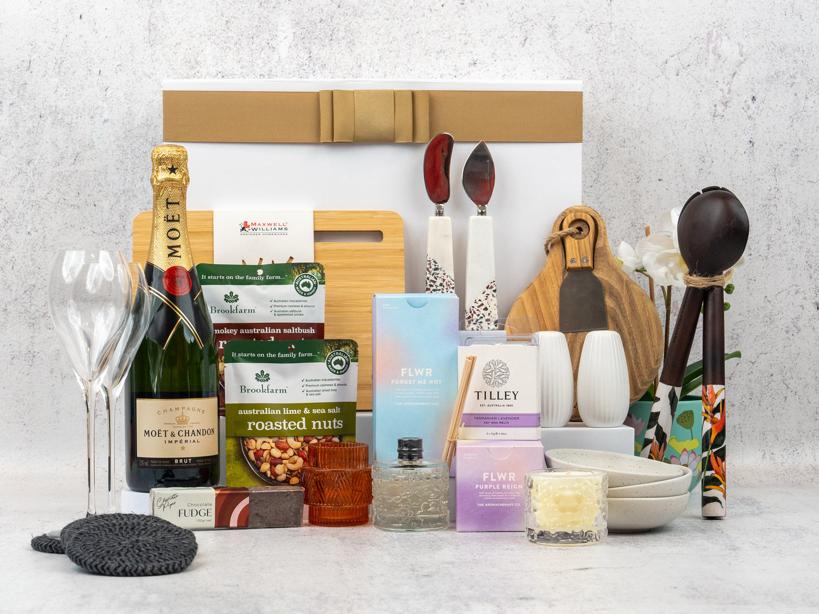 The Hamper Boutique Co’s Ultimate Homeware Gift Hamper features Moët & Chandon champagne, flutes, snacks, candles, fudge, nuts, utensils, a plant, bowls and cutting boards—beautifully arranged in front of a gift box.
