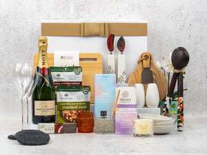 The Hamper Boutique Co’s Ultimate Homeware Gift Hamper features Moët & Chandon champagne, flutes, snacks, candles, fudge, nuts, utensils, a plant, bowls and cutting boards—beautifully arranged in front of a gift box.