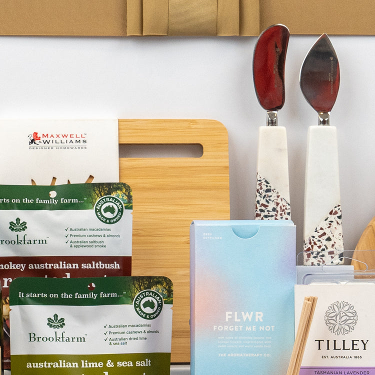 The Hamper Boutique Co's Ultimate Homeware Gift Hamper features a wooden cutting board, red-handled ceramic cheese knives, Brookfarm nut snacks, FLWR tissues, and Tilley Tasmanian lavender diffuser sticks.