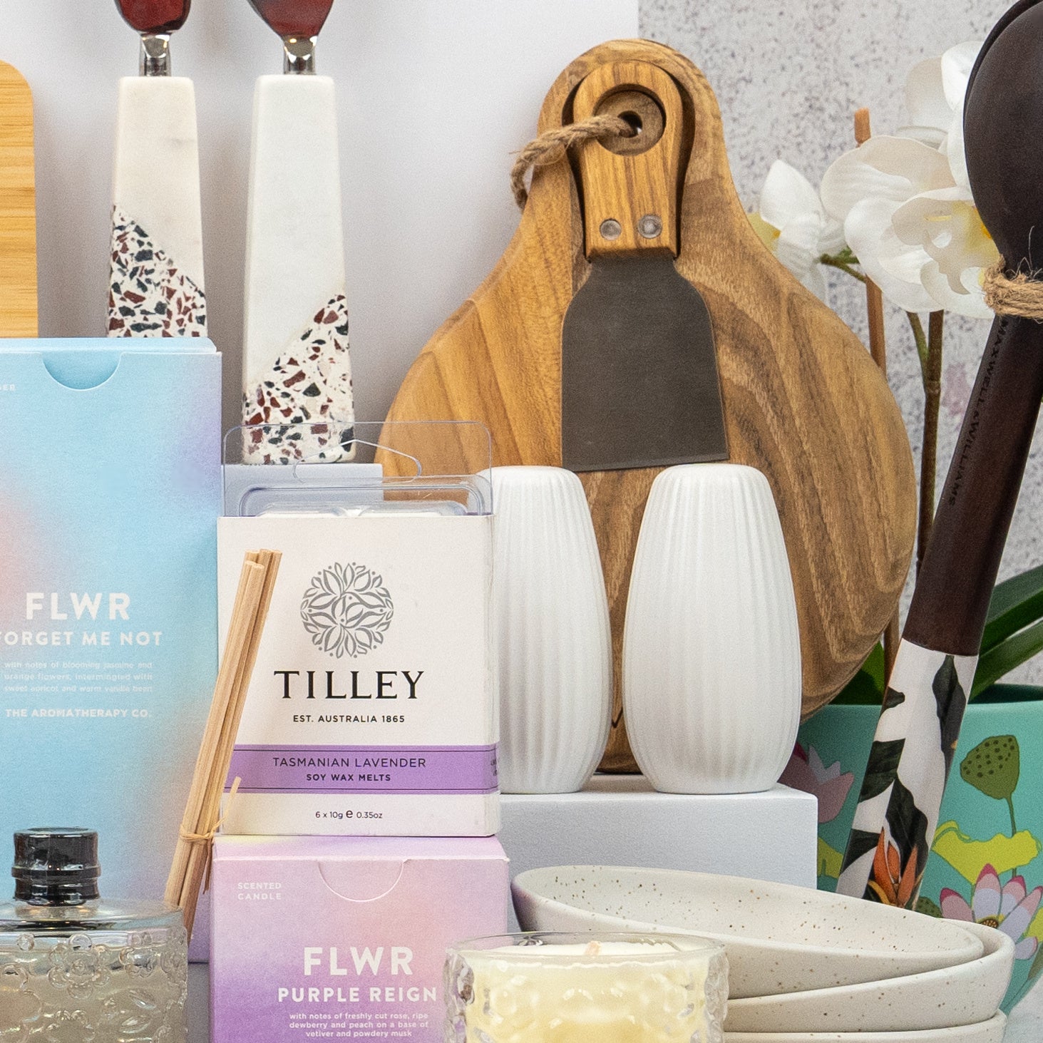 The Hamper Boutique Co's Ultimate Homeware Gift Hamper features chopping boards, salt and pepper shakers, bowls, a candle, reed diffuser, boxed soaps, utensils, and white artificial orchids arranged beautifully together.