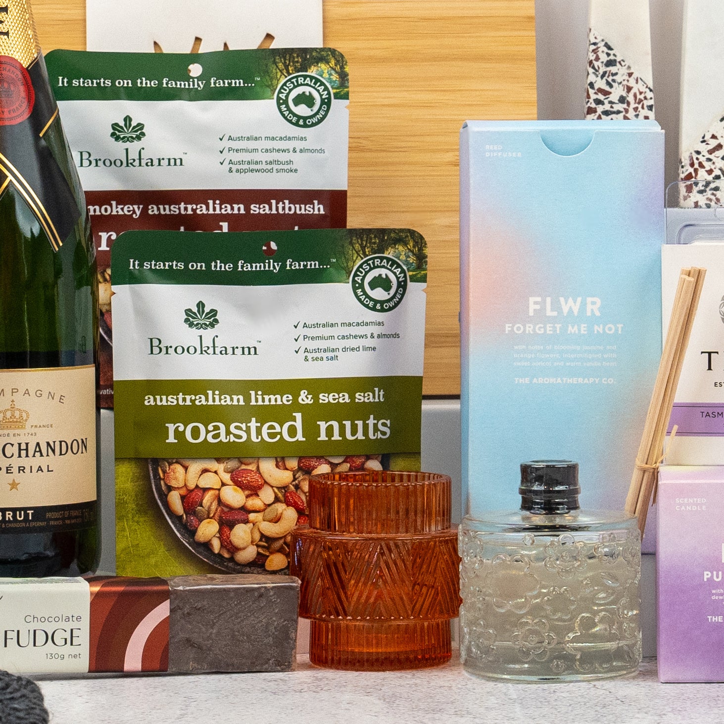 A close-up of The Hamper Boutique Co's Ultimate Homeware Gift Hamper, featuring Brookfarm roasted nuts, a FLWR Forget Me Not box, an amber candle, a textured bottle, and part of a Moët & Chandon champagne box.