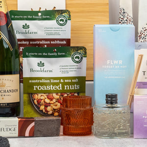 A close-up of The Hamper Boutique Co's Ultimate Homeware Gift Hamper, featuring Brookfarm roasted nuts, a FLWR Forget Me Not box, an amber candle, a textured bottle, and part of a Moët & Chandon champagne box.