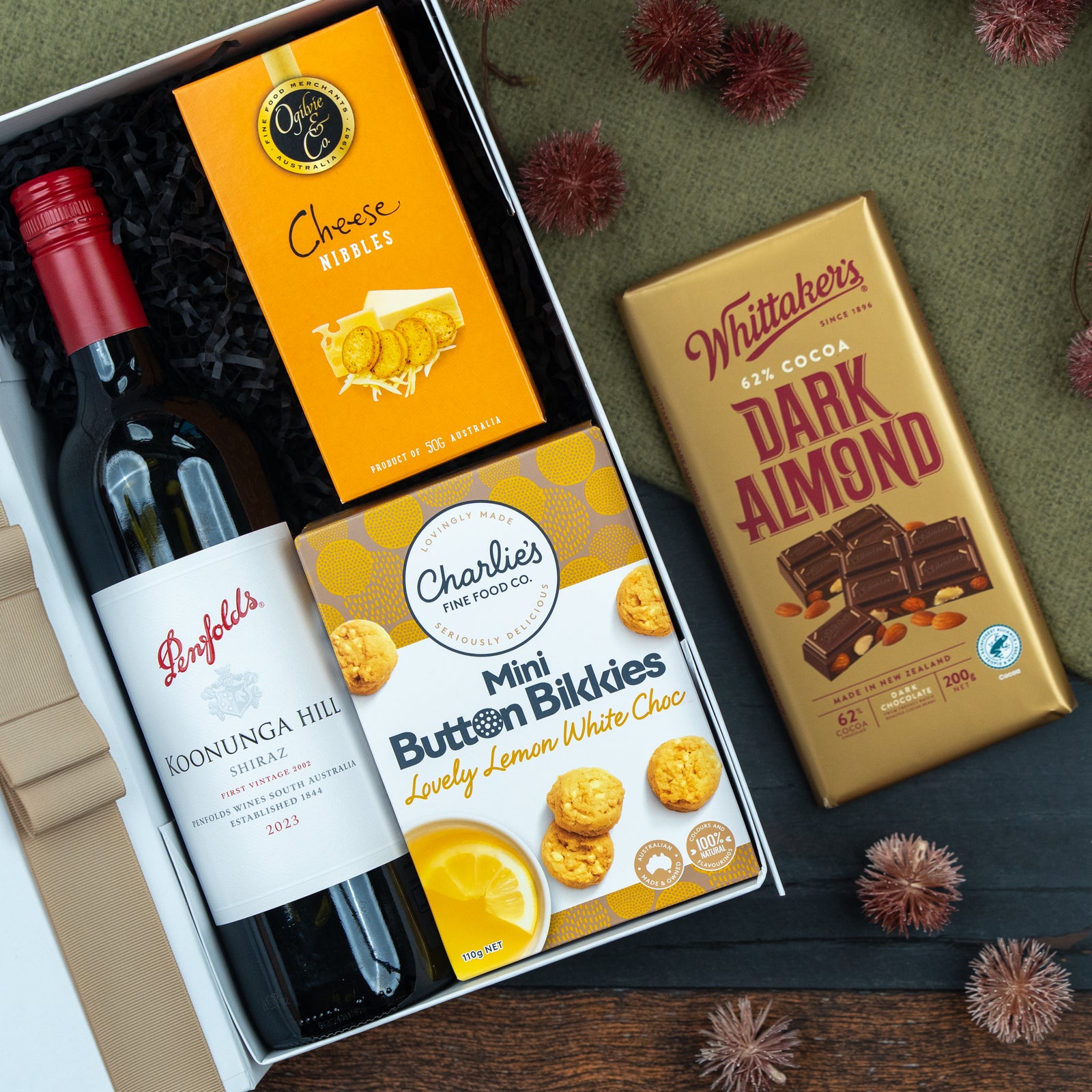 The Hamper Boutique Co’s Classic Delights Gift Hamper features Penfolds red wine, Cheese Nibbles crackers, Charlie’s Mini Button Bikkies, and a Whittaker’s Dark Almond chocolate bar, presented with decorative elements on a green surface.