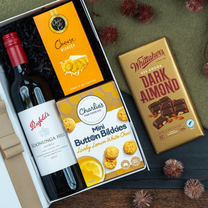 The Hamper Boutique Co’s Classic Delights Gift Hamper features Penfolds red wine, Cheese Nibbles crackers, Charlie’s Mini Button Bikkies, and a Whittaker’s Dark Almond chocolate bar, presented with decorative elements on a green surface.