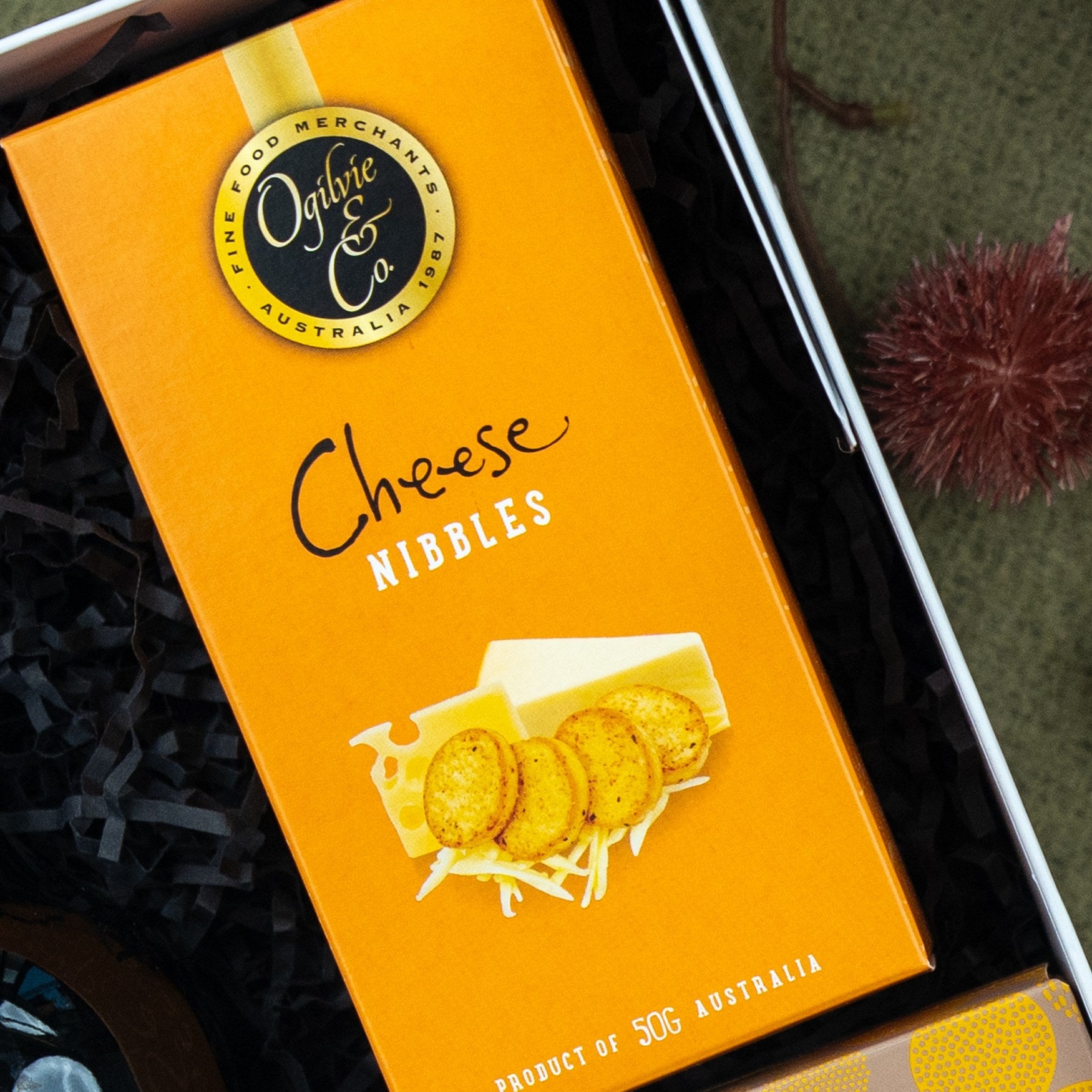 A Classic Delights Gift Hamper by The Hamper Boutique Co includes a yellow box of cheese nibbles with crackers and cheese imagery, nestled in black shredded paper. The 50g box is a product of Australia.