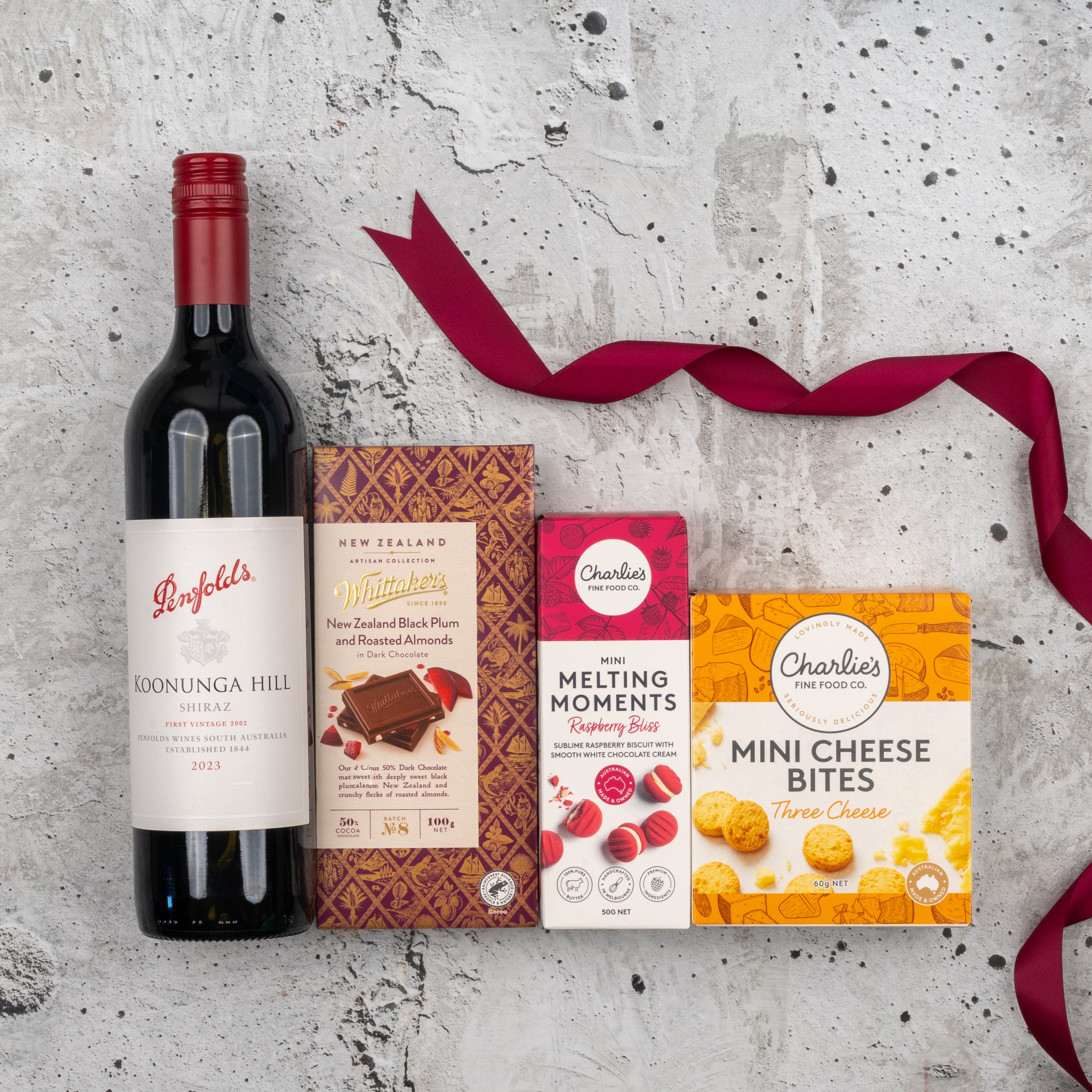 The Hamper Boutique Co Penfolds Shiraz Tasting Hamper, featuring wine and gourmet treats, is displayed on a gray textured surface with a red ribbon.