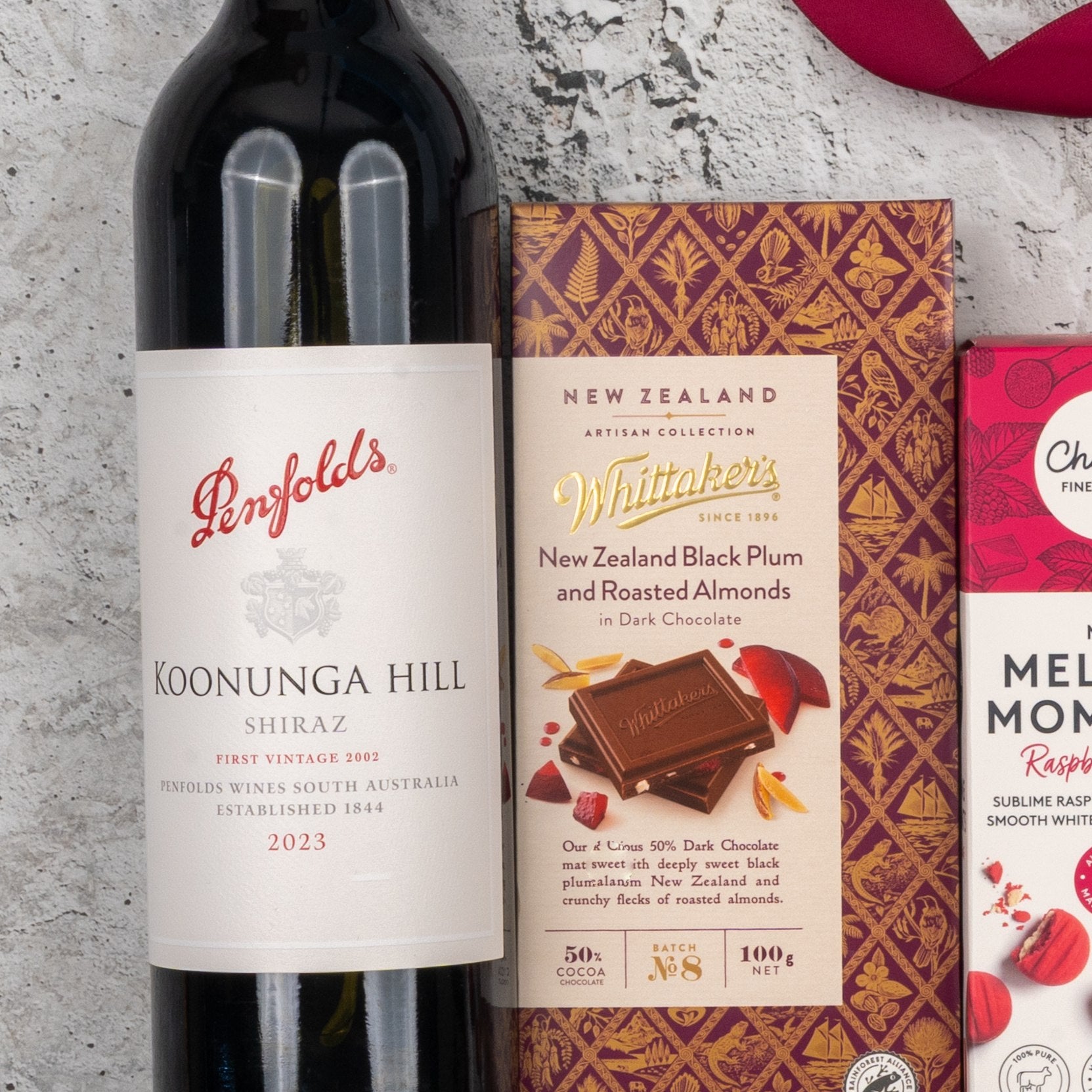 A Penfolds Shiraz Tasting Hamper from The Hamper Boutique Co is shown on a light textured surface, with the hamper featuring wine and boxes of gourmet dark chocolate, including Whittakers New Zealand Black Plum and Roasted Almonds.