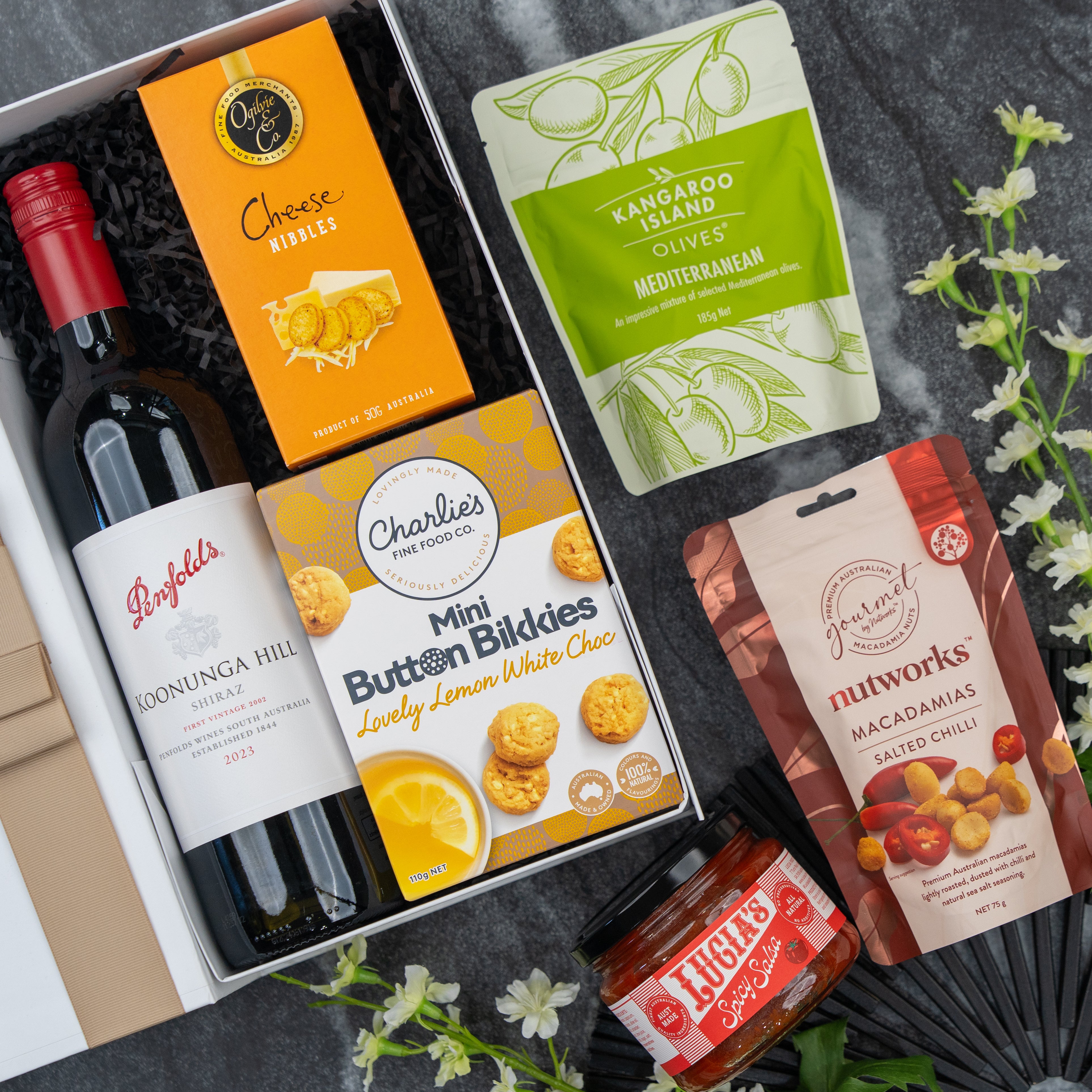 The Hamper Boutique Co’s Gourmet Journey Gift Hamper includes Penfolds red wine, cheese nibbles, lemon white choc cookies, Mediterranean olives, spiced macadamias & chili jam, beautifully presented with white flowers.