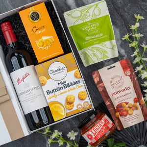 The Hamper Boutique Co’s Gourmet Journey Gift Hamper includes Penfolds red wine, cheese nibbles, lemon white choc cookies, Mediterranean olives, spiced macadamias & chili jam, beautifully presented with white flowers.