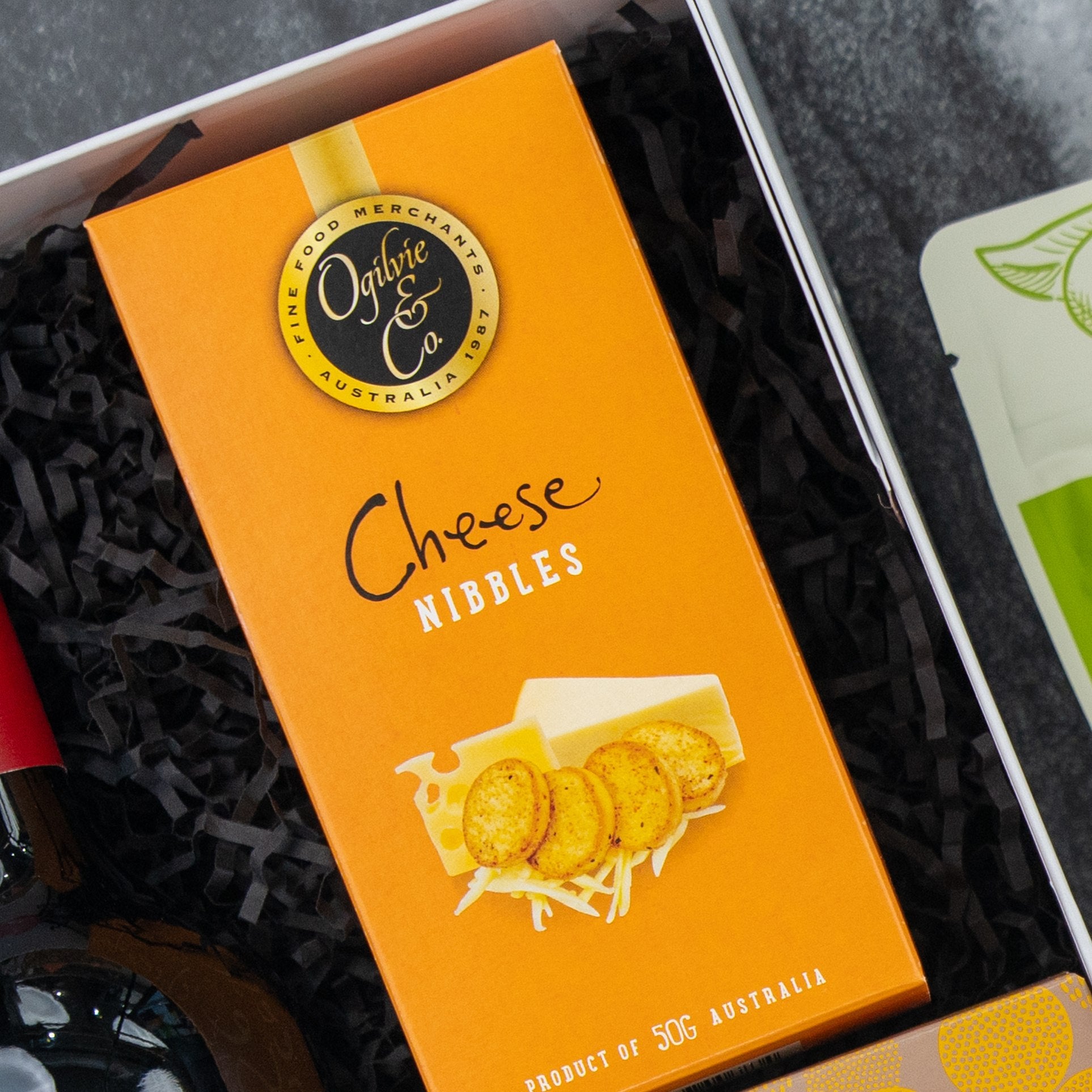 A Gourmet Journey Gift Hamper by The Hamper Boutique Co features an orange box of cheese nibbles with a cheese and crackers image, presented in a gift box lined with black shredded paper. Product of Australia.