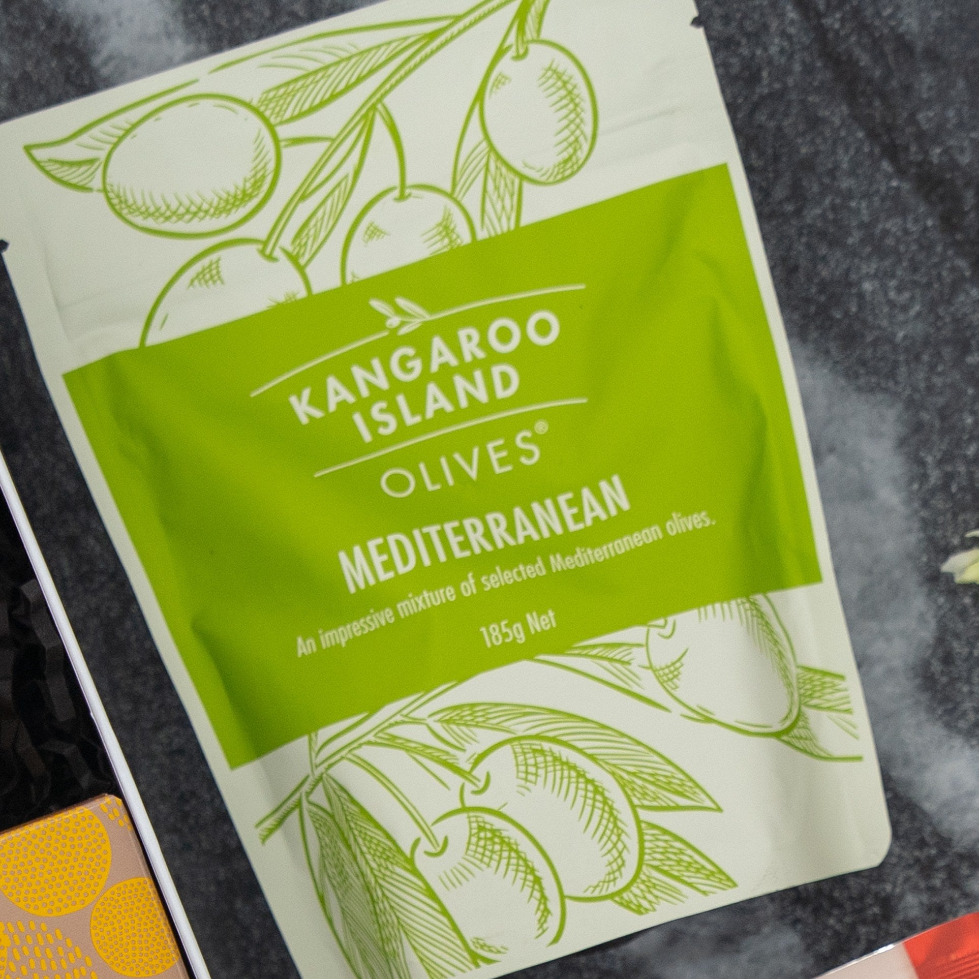 A green and white bag from The Hamper Boutique Co’s Gourmet Journey Gift Hamper rests on dark marble. Its packaging, decorated with olive illustrations, describes it as a mix of Mediterranean olives, 185g net weight.