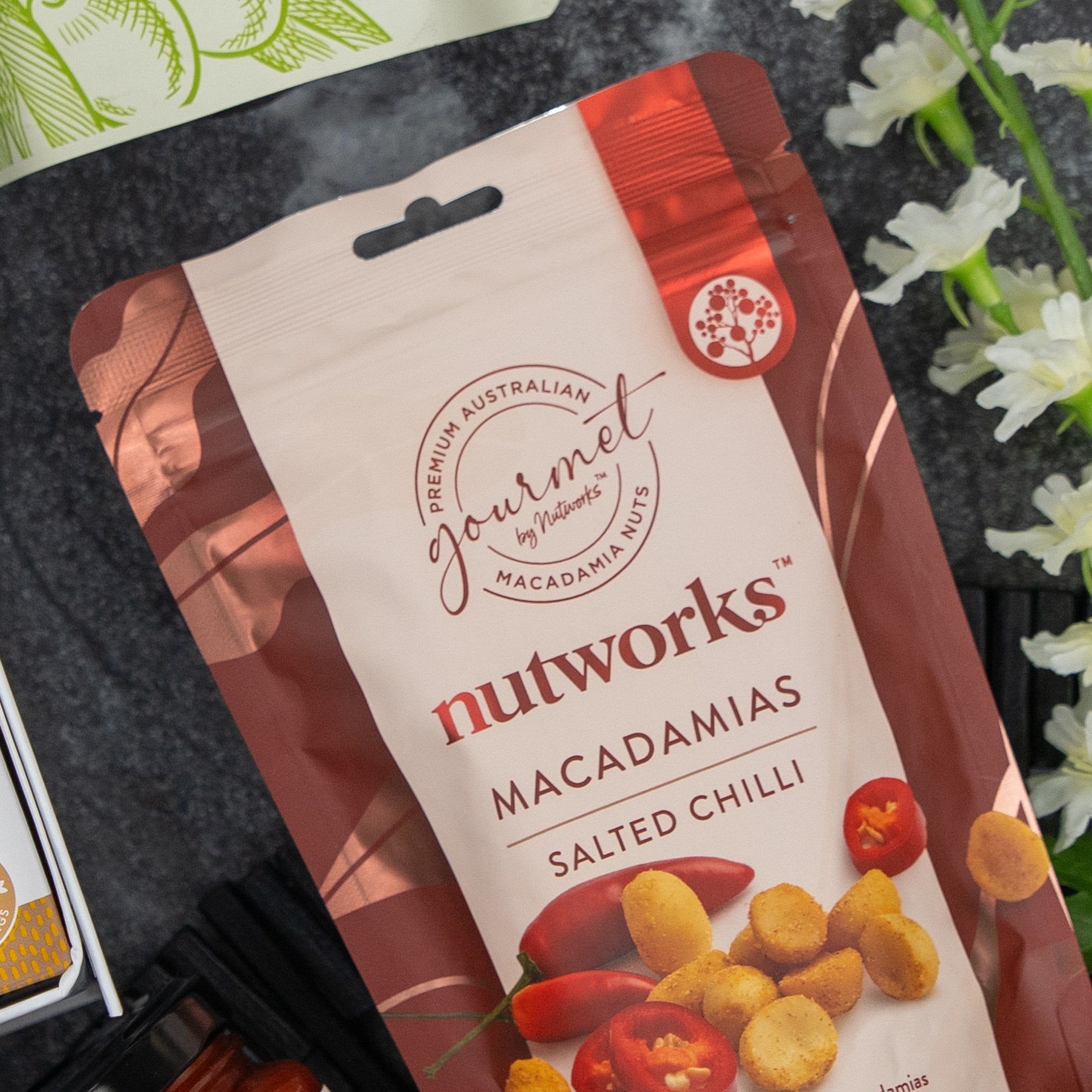 A Gourmet Journey Gift Hamper by The Hamper Boutique Co sits on a table among flowers and other items. Its packaging is red and beige, featuring images of macadamia nuts and chilli.