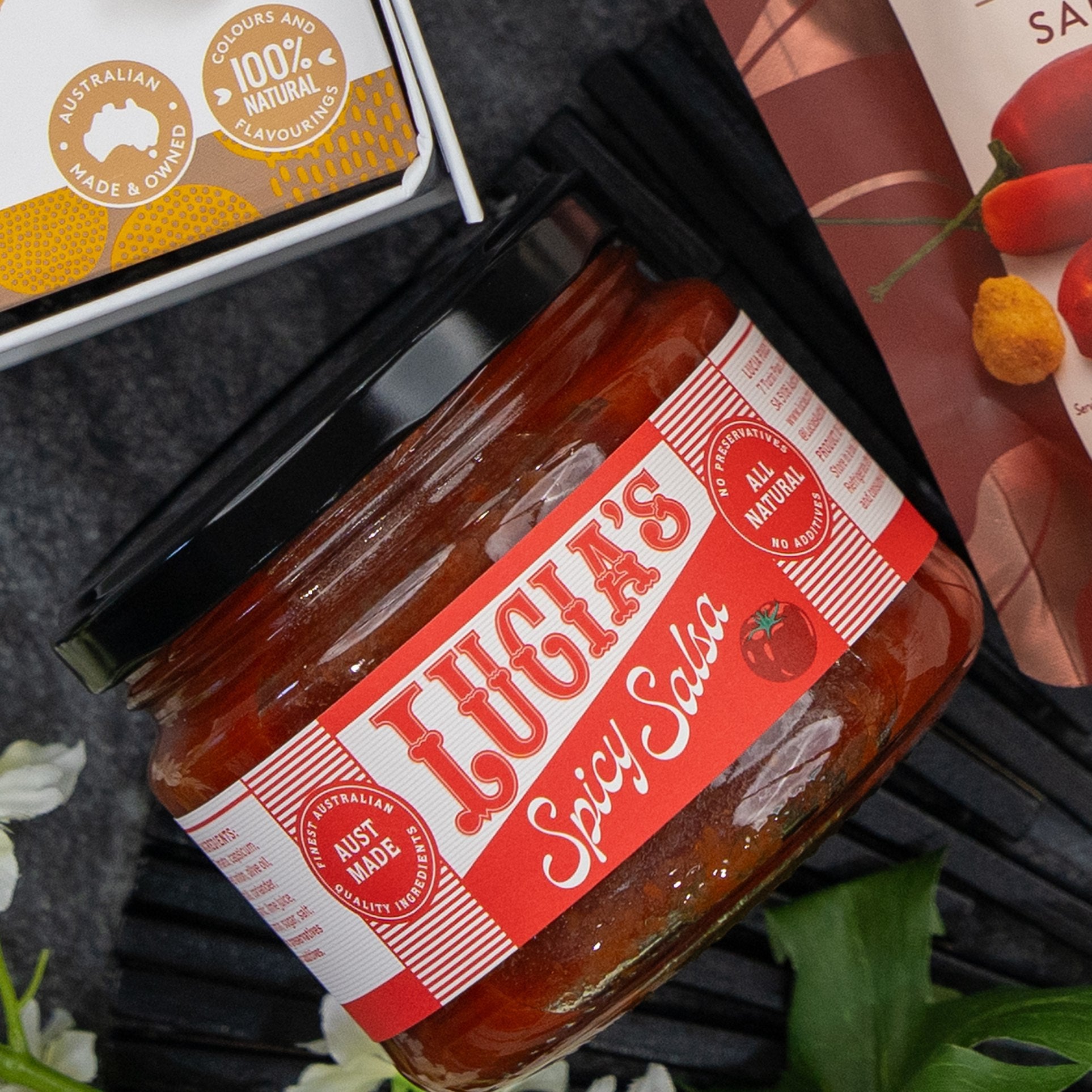 A Gourmet Journey Gift Hamper by The Hamper Boutique Co is displayed, featuring a jar with a red label emphasizing its Australian-made and all-natural qualities. Other food packaging can be seen nearby.