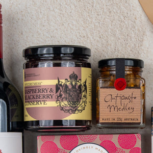 A close-up of The Hamper Boutique Co Taste Explorer Gift Hamper, featuring jars of raspberry & blackberry conserve and antipasto medley, part of a wine bottle, and a pink patterned box arranged on a light surface.
