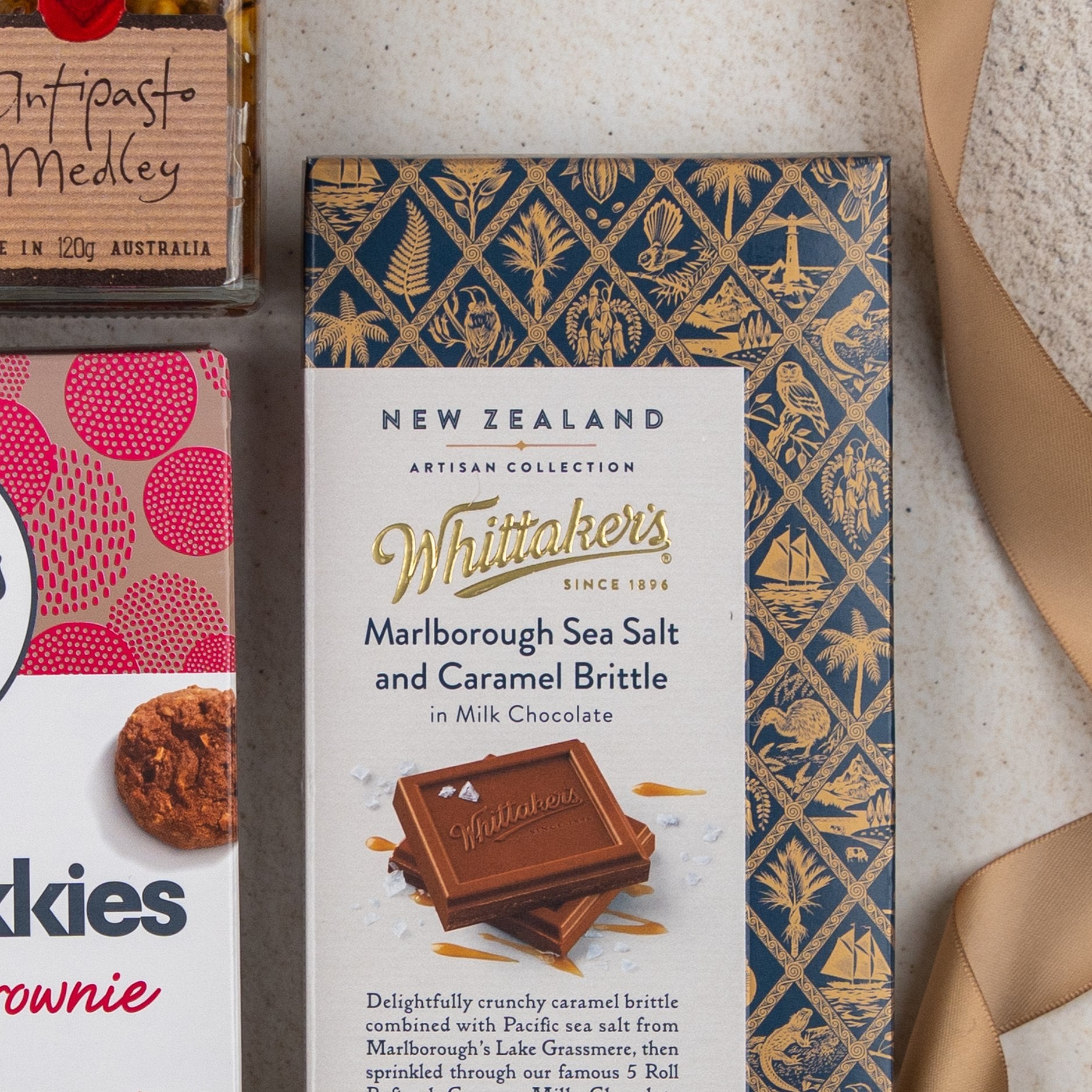 A close-up of The Hamper Boutique Co Taste Explorer Gift Hamper, featuring gourmet treats like Marlborough Sea Salt & Caramel Brittle chocolate, antipasto medley, cookies, and a gold satin ribbon on a light surface.