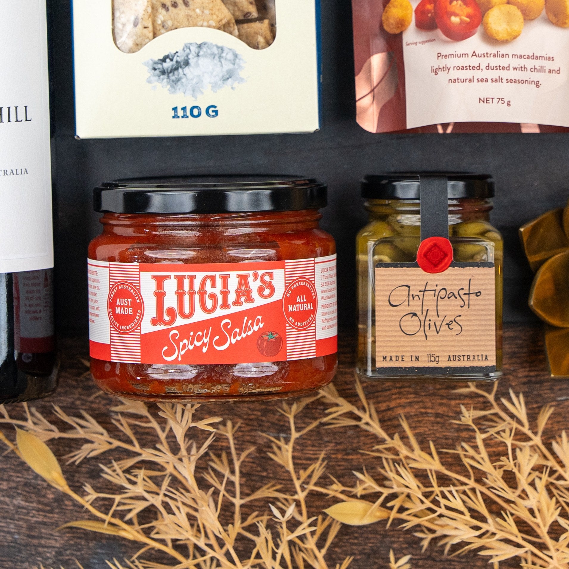 The Hamper Boutique Co's Savoury Selection Gift Hamper features items like antipasto olives and spicy salsa, beautifully arranged with a wine bottle and crackers, accented by dried leaves.