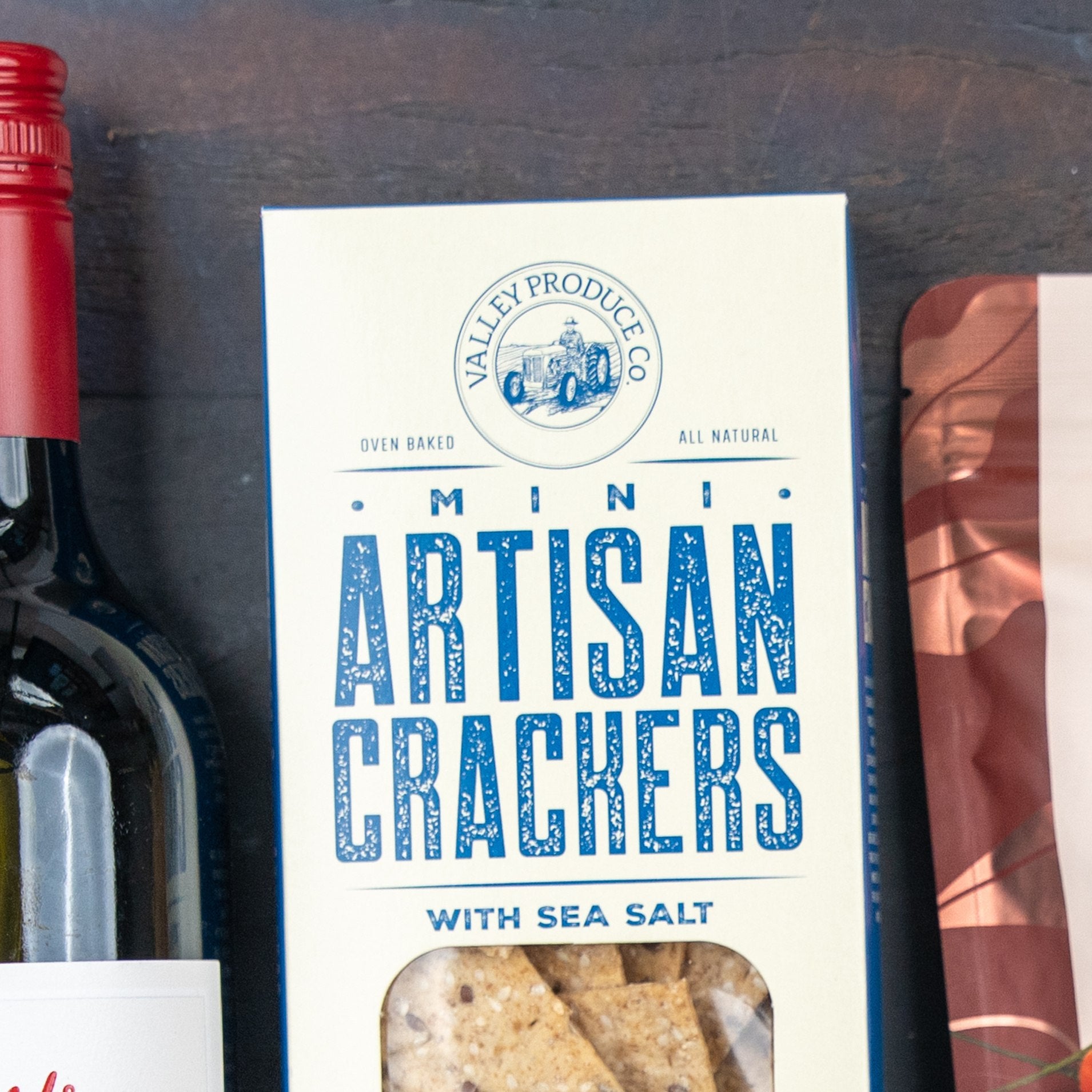 A close-up of The Hamper Boutique Co Savoury Selection Gift Hamper beside a bottle of red wine and part of another package on a dark surface.